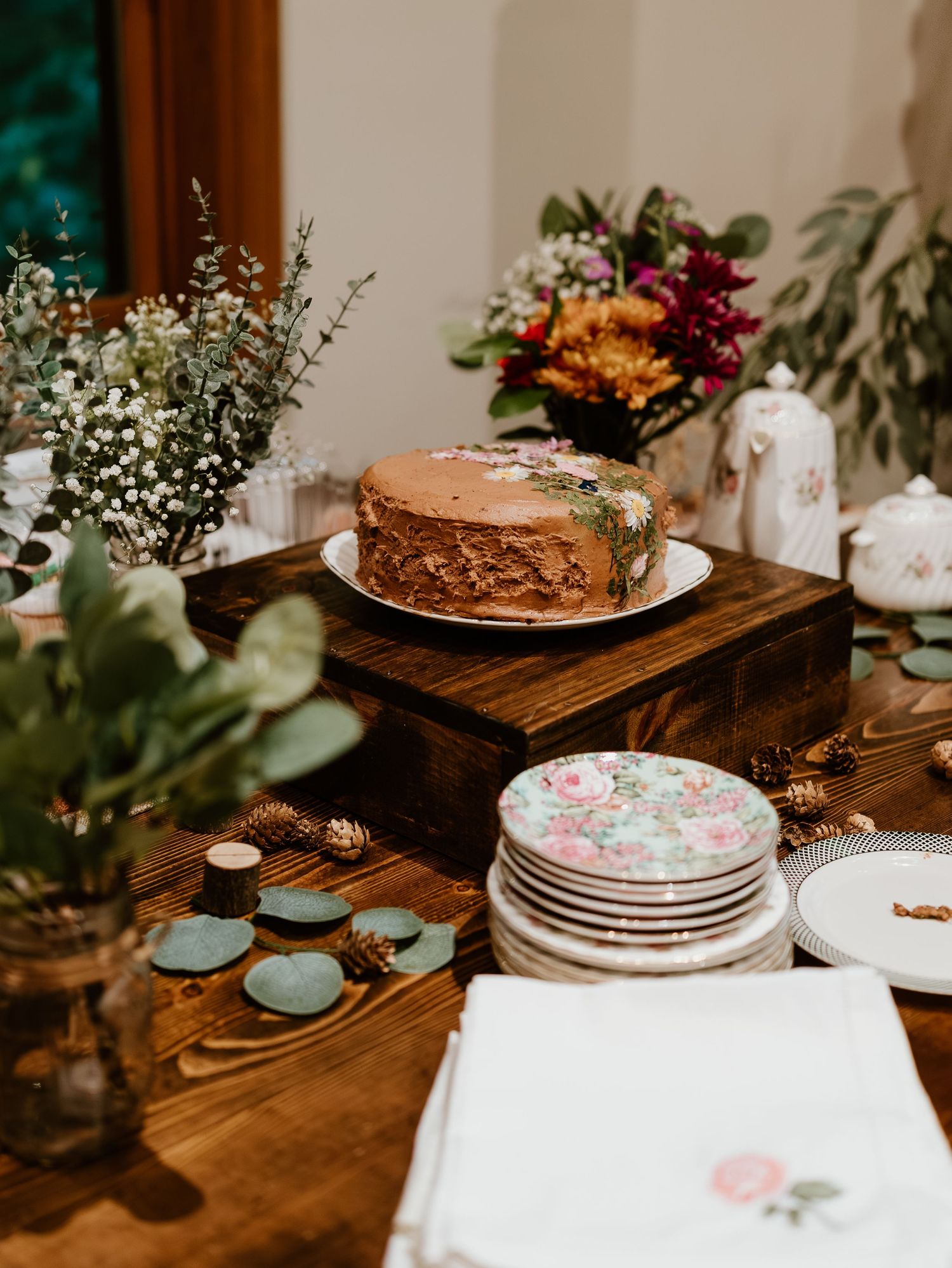 Rustic wedding dessert table with chocolate and floral decorated cakes on wooden surfaces with greenery accents.