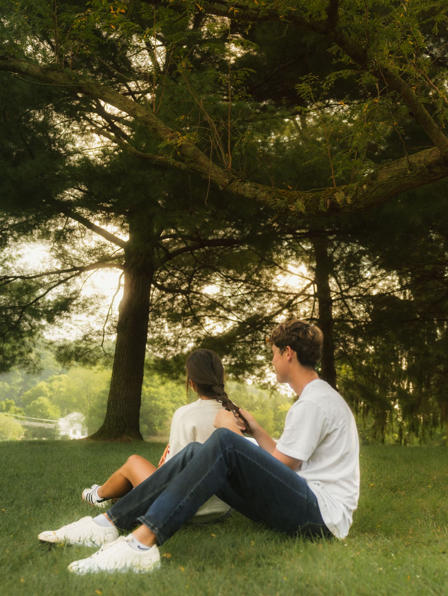 A couple sits on grass under large trees in a park, enjoying a peaceful sunset together.
