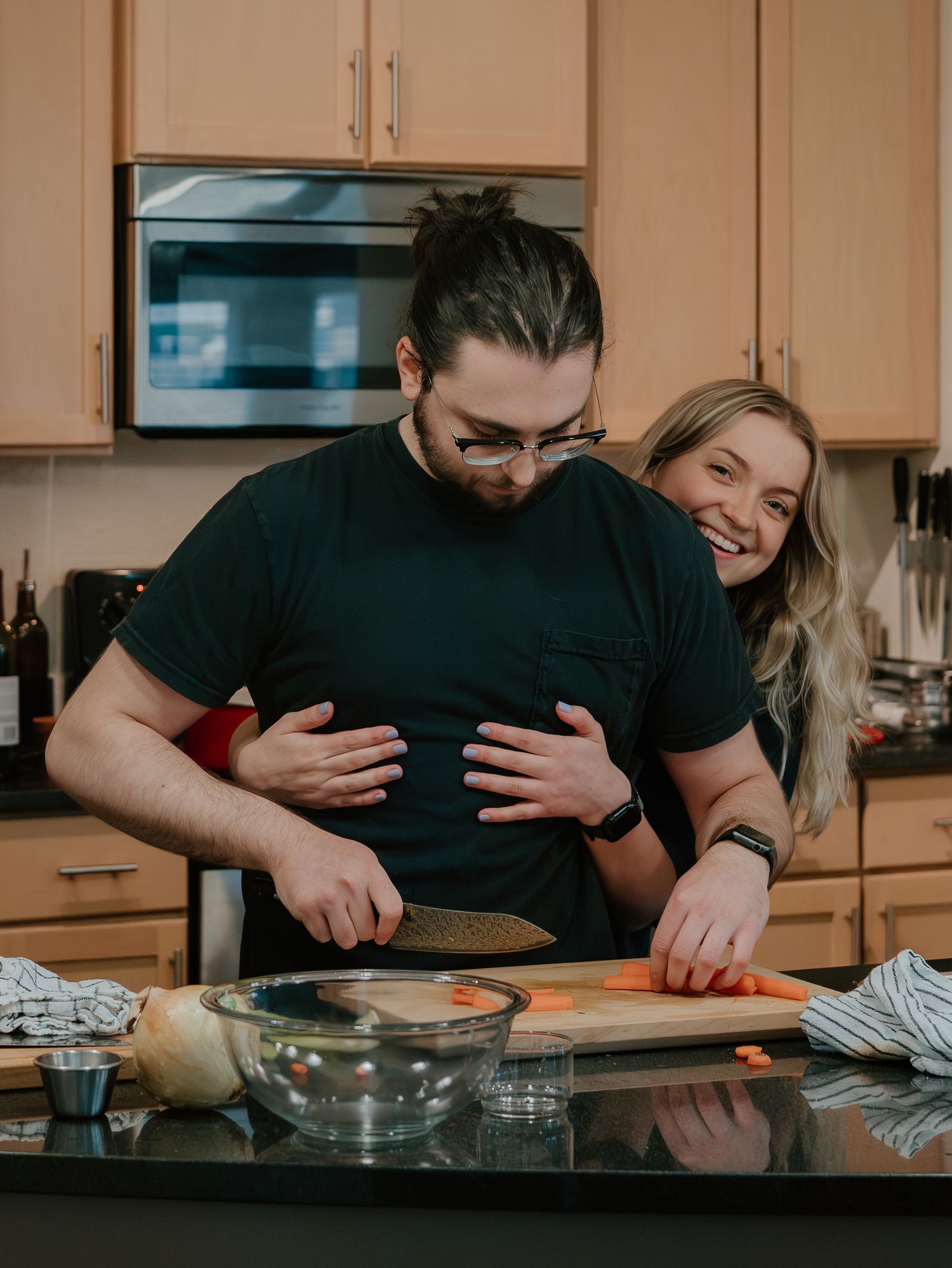 A couple embraces while baking together in a modern kitchen with wooden cabinets and stainless steel appliances.