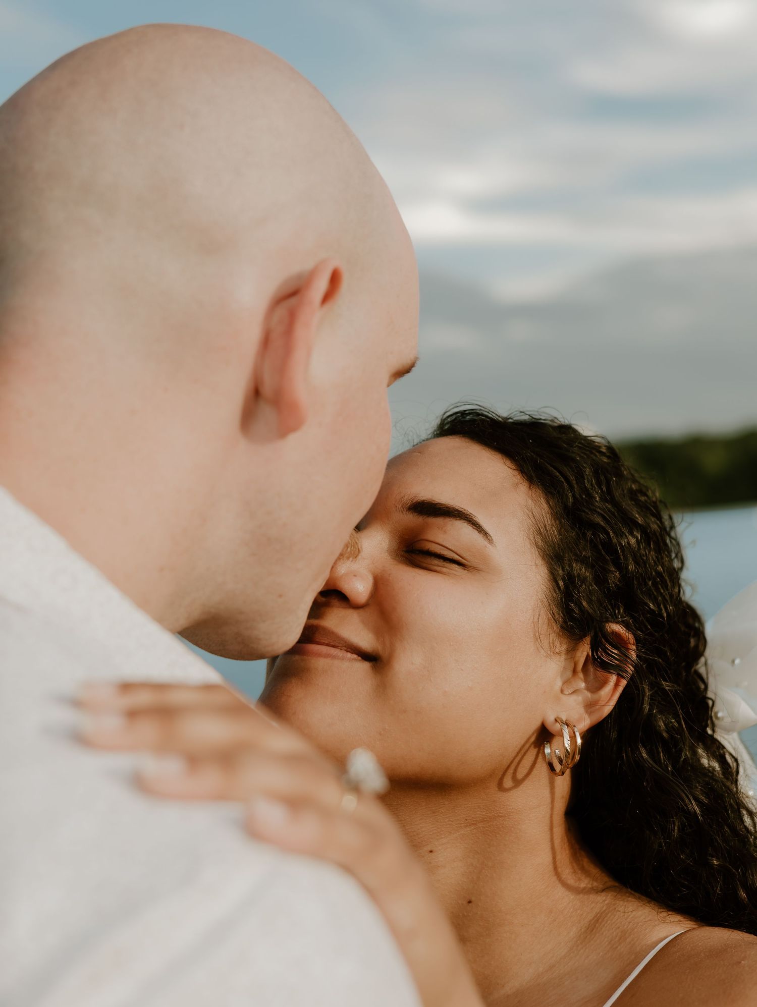 A romantic couple sharing an intimate kiss outdoors with a cloudy sky backdrop.