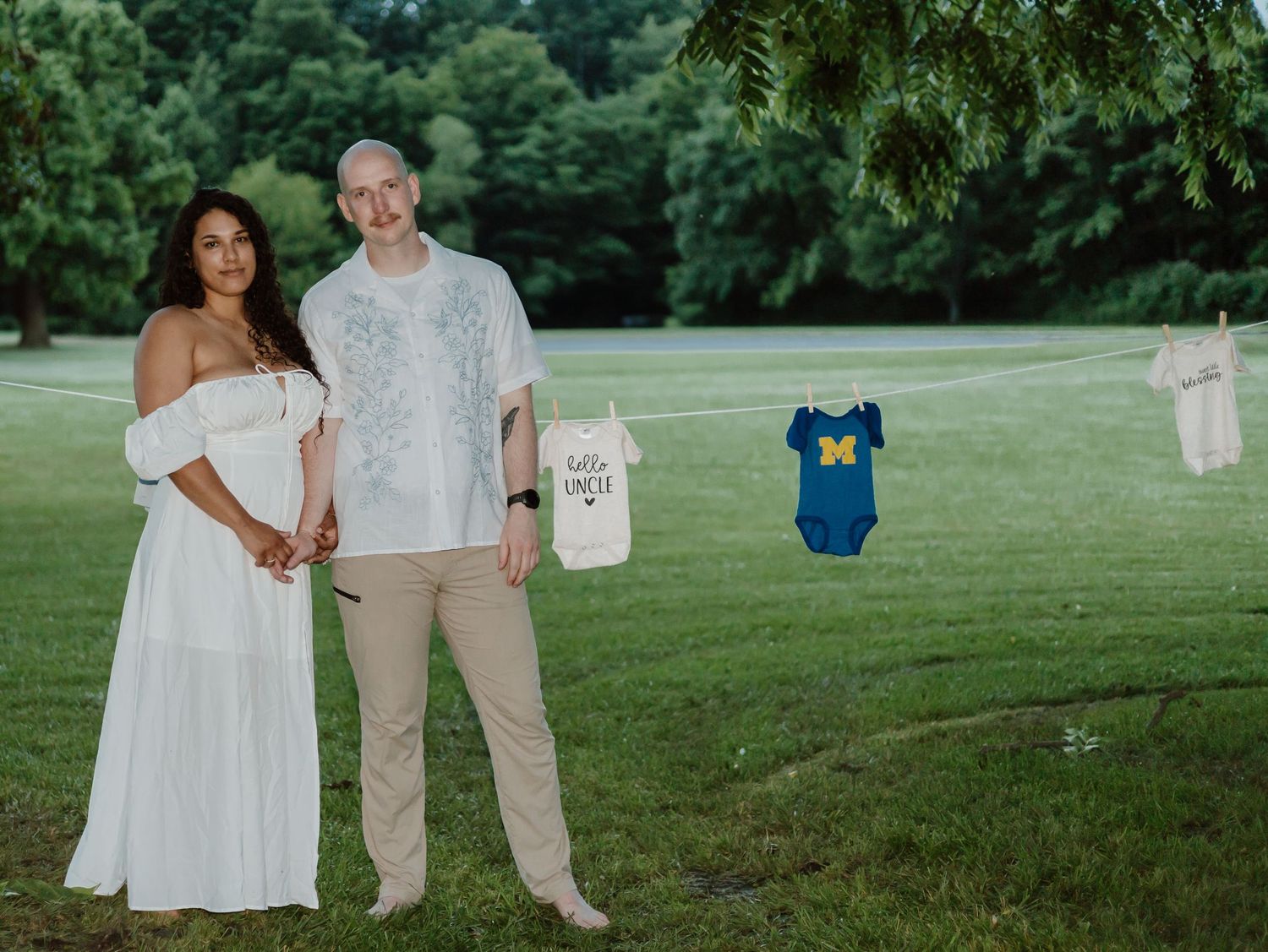 A couple poses together on a grassy lawn holding up baby clothes and onesies during a gender reveal announcement.
