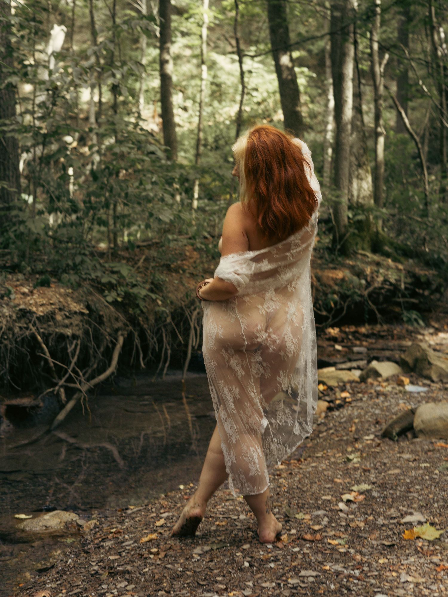 A person wrapped in a sheer fabric stands beside a stream in a misty forest setting.