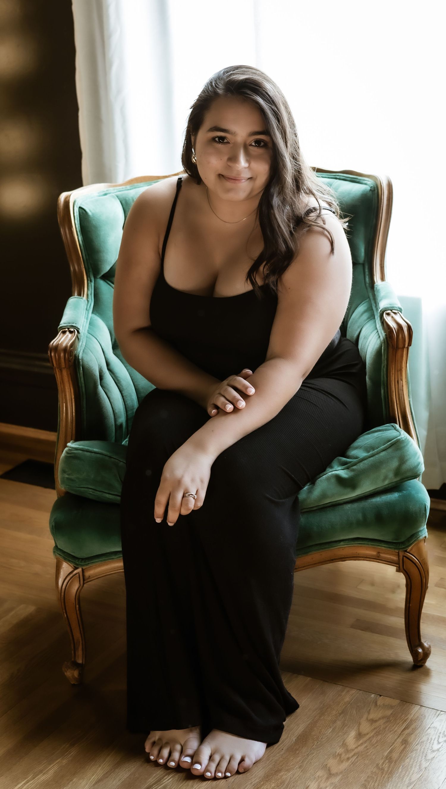 Someone in a black outfit sits relaxed in a vintage mint green velvet armchair on hardwood floors.