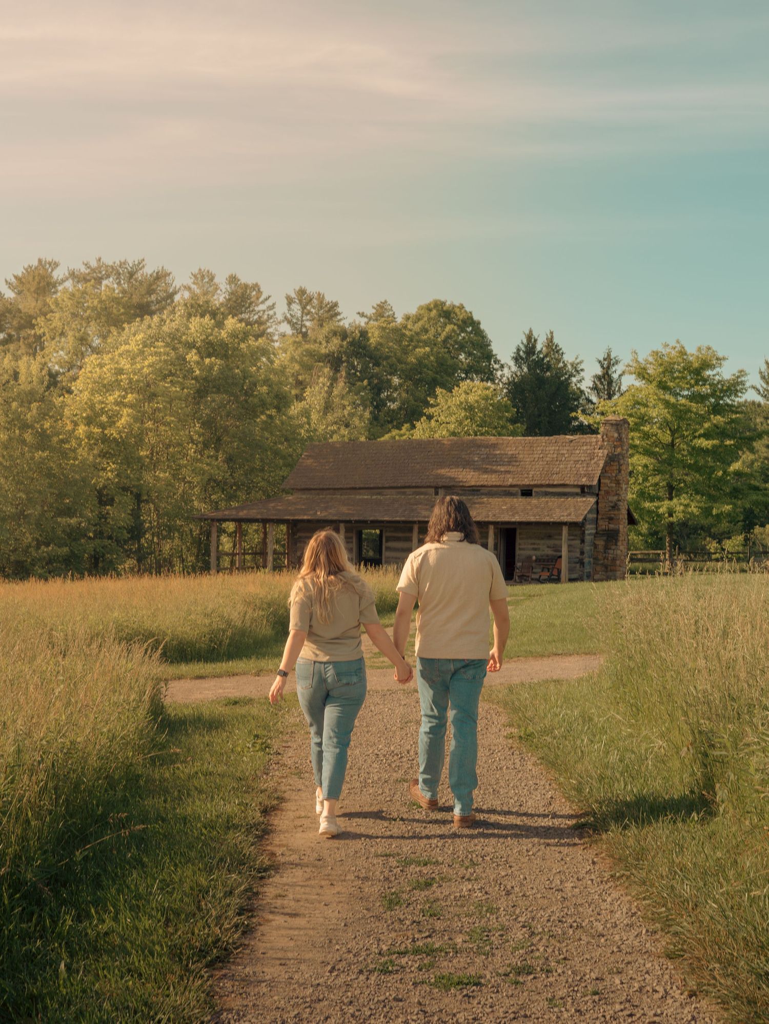 A couple walks hand in hand down a dirt path towards a rustic cabin surrounded by autumn trees.