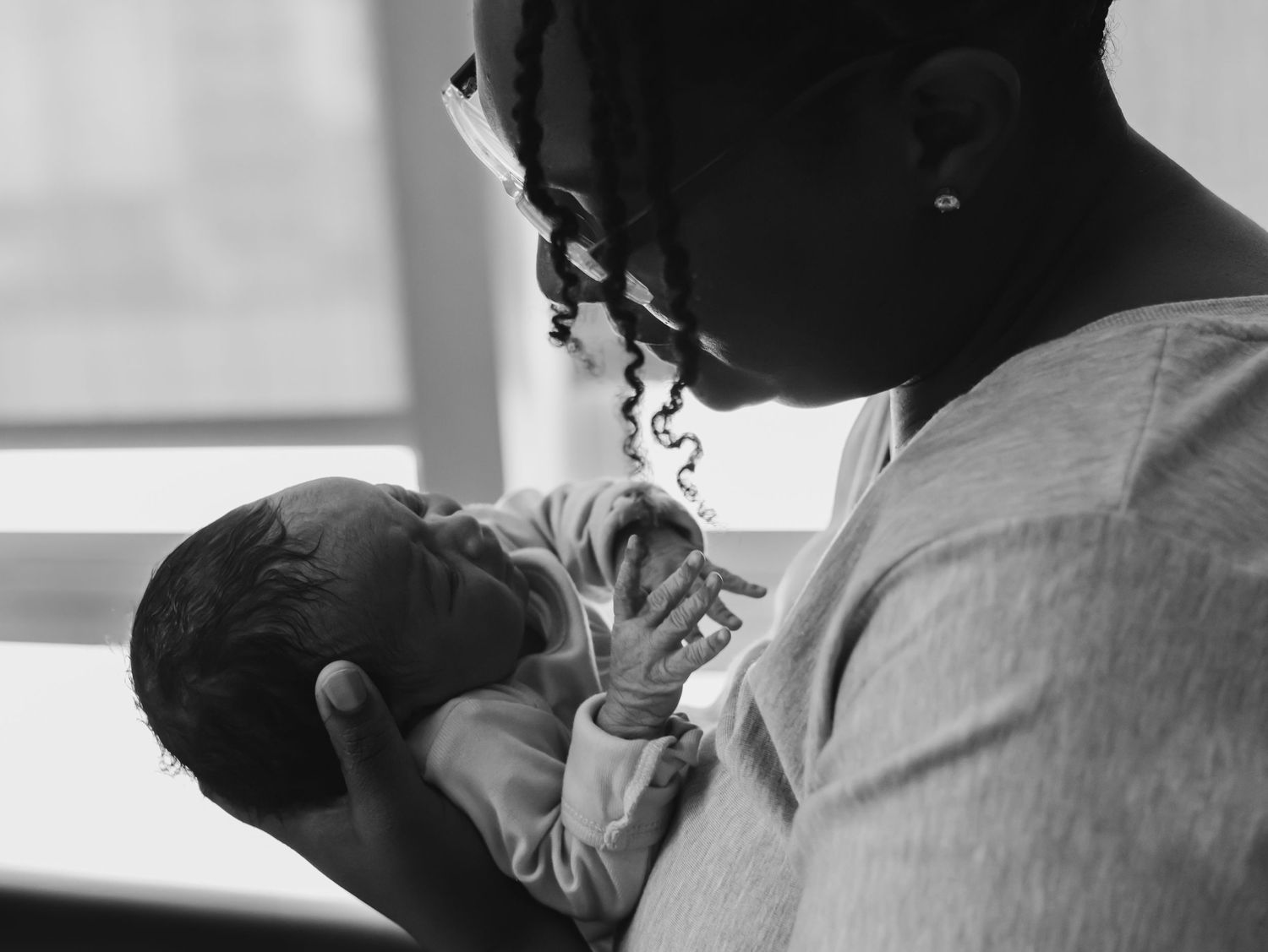 A tender black and white photograph captures a parent lovingly cradling their newborn baby in soft window light.