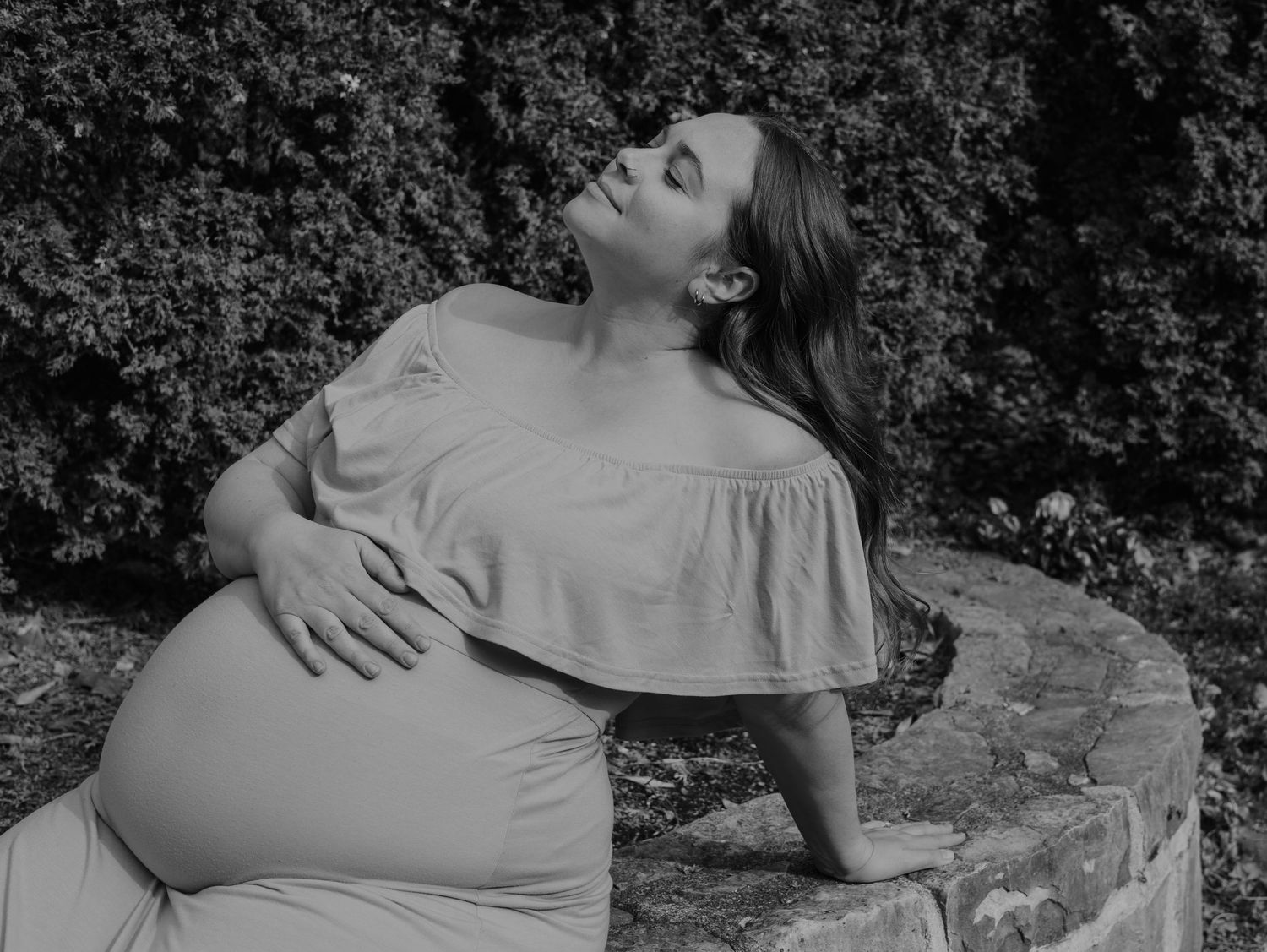 A pregnant woman in a ruffled dress poses gracefully against stone steps and foliage in a black and white portrait.