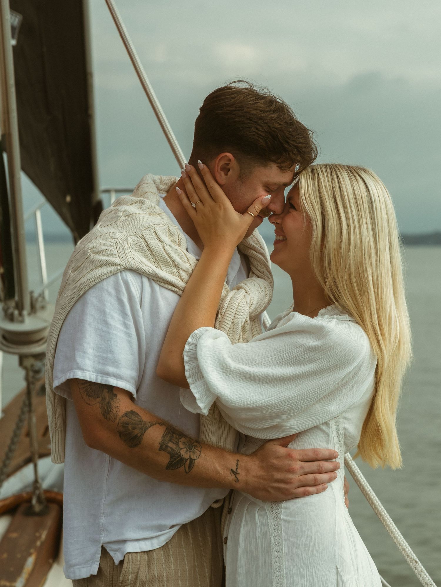 A couple in white outfits share a romantic moment on a sailboat, with the man's tattooed arms embracing the blonde woman.
