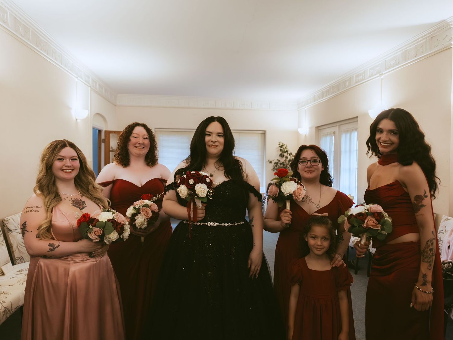 Bride in black gown with bridesmaids in burgundy and mauve dresses holding floral bouquets at wedding.