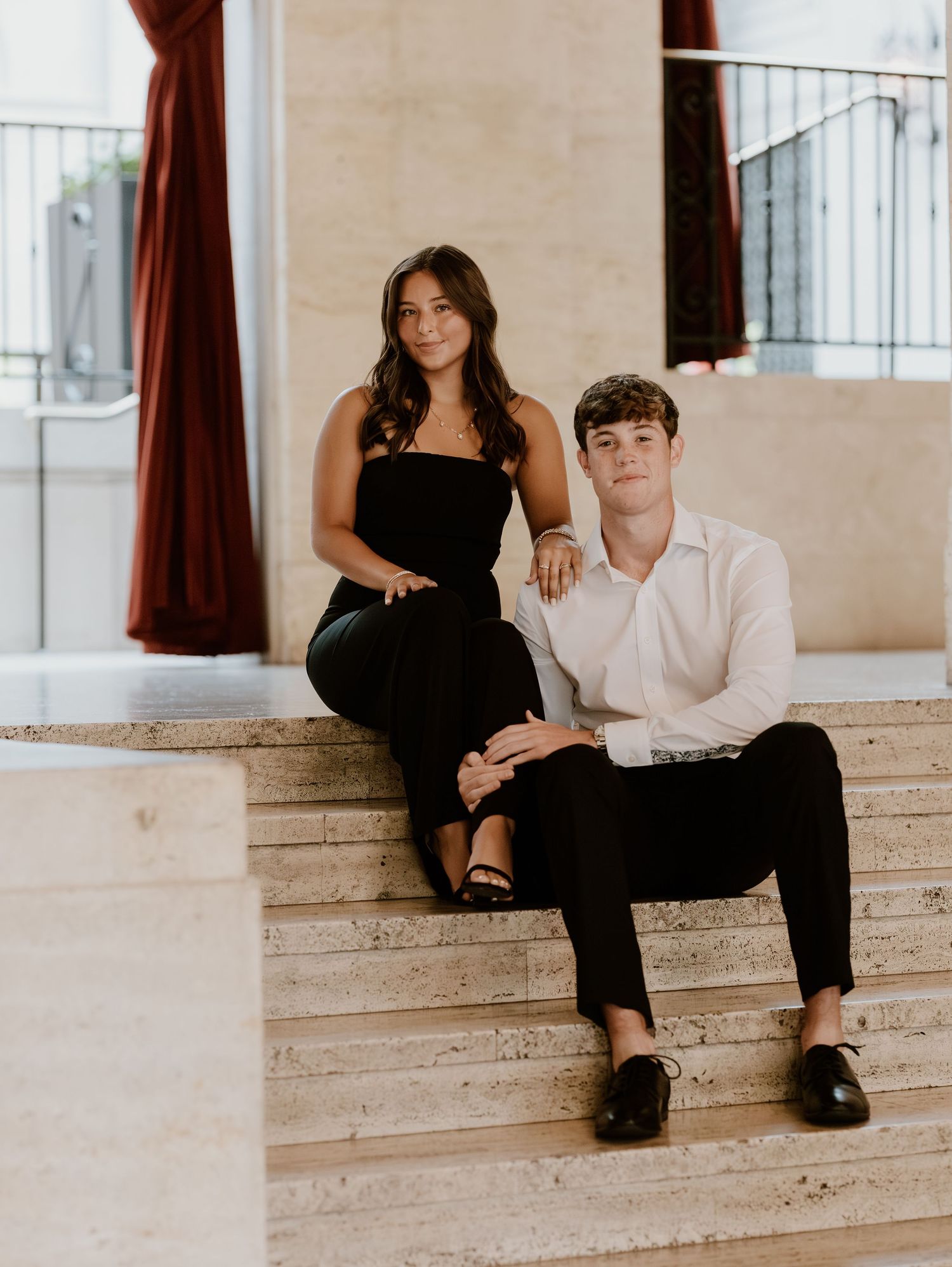 A couple in black and white outfits sits casually on stone steps in an elegant indoor setting.