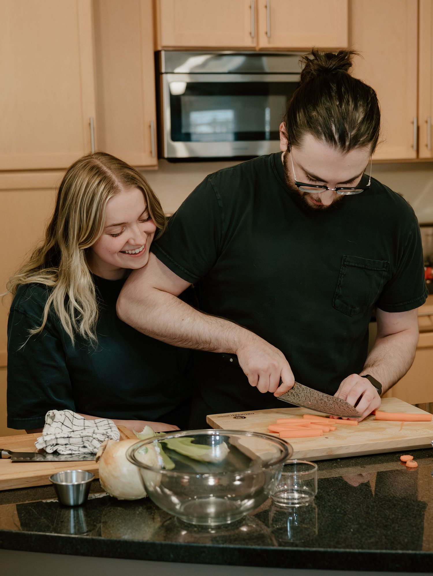 A couple in a kitchen works together to prepare food on a dark countertop with cooking ingredients and utensils.