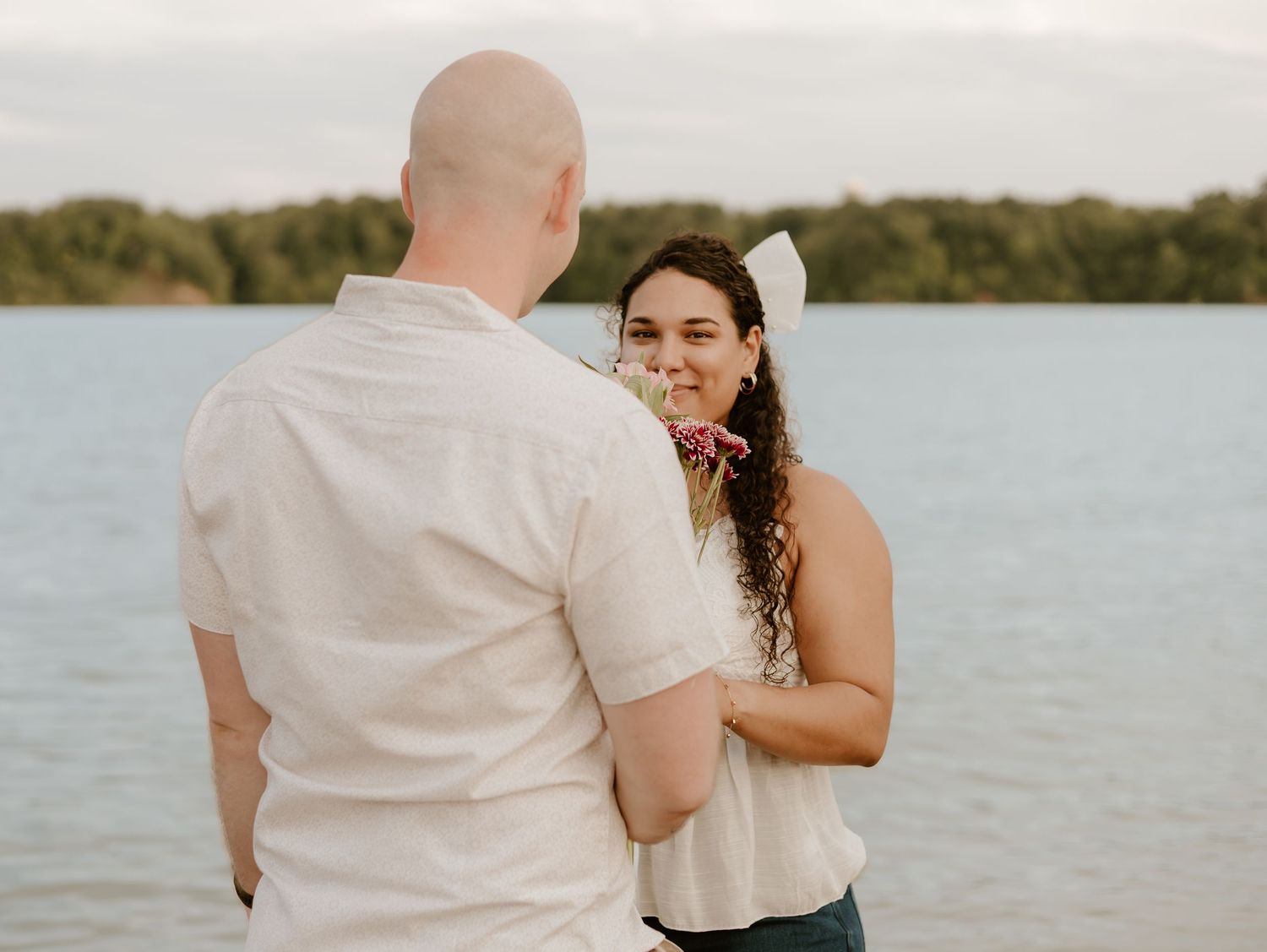 A couple in white embraces by a serene lakeside during a romantic outdoor photoshoot.