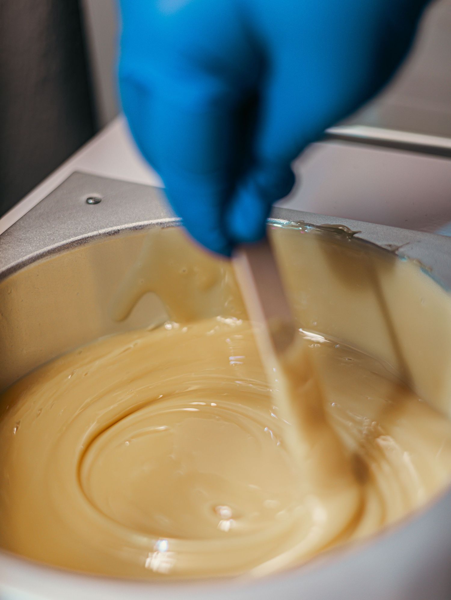 A hand wearing blue gloves stirs smooth yellow custard in a white bowl with a metal utensil.