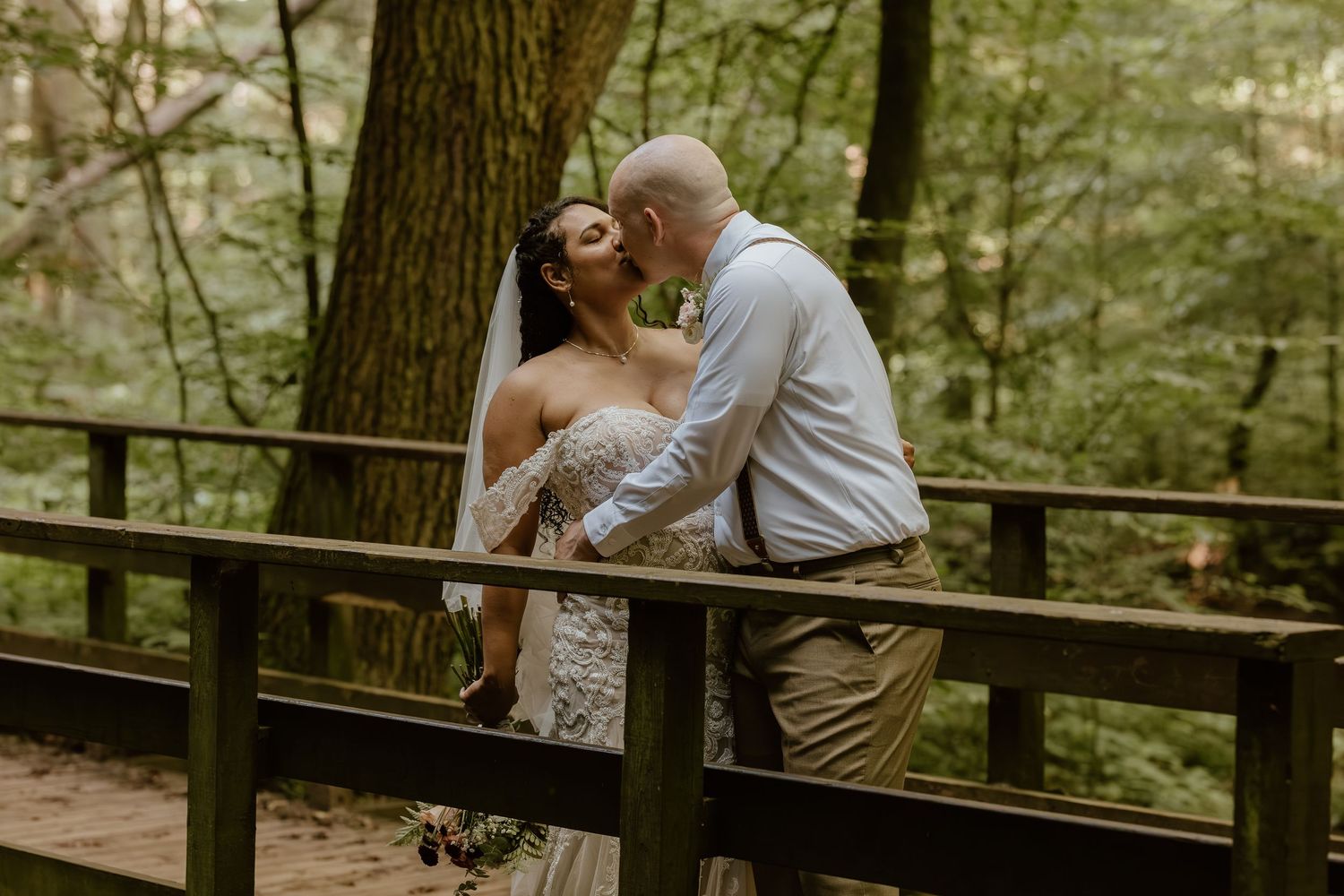Newlyweds share a romantic moment on a wooden bridge surrounded by lush forest greenery.