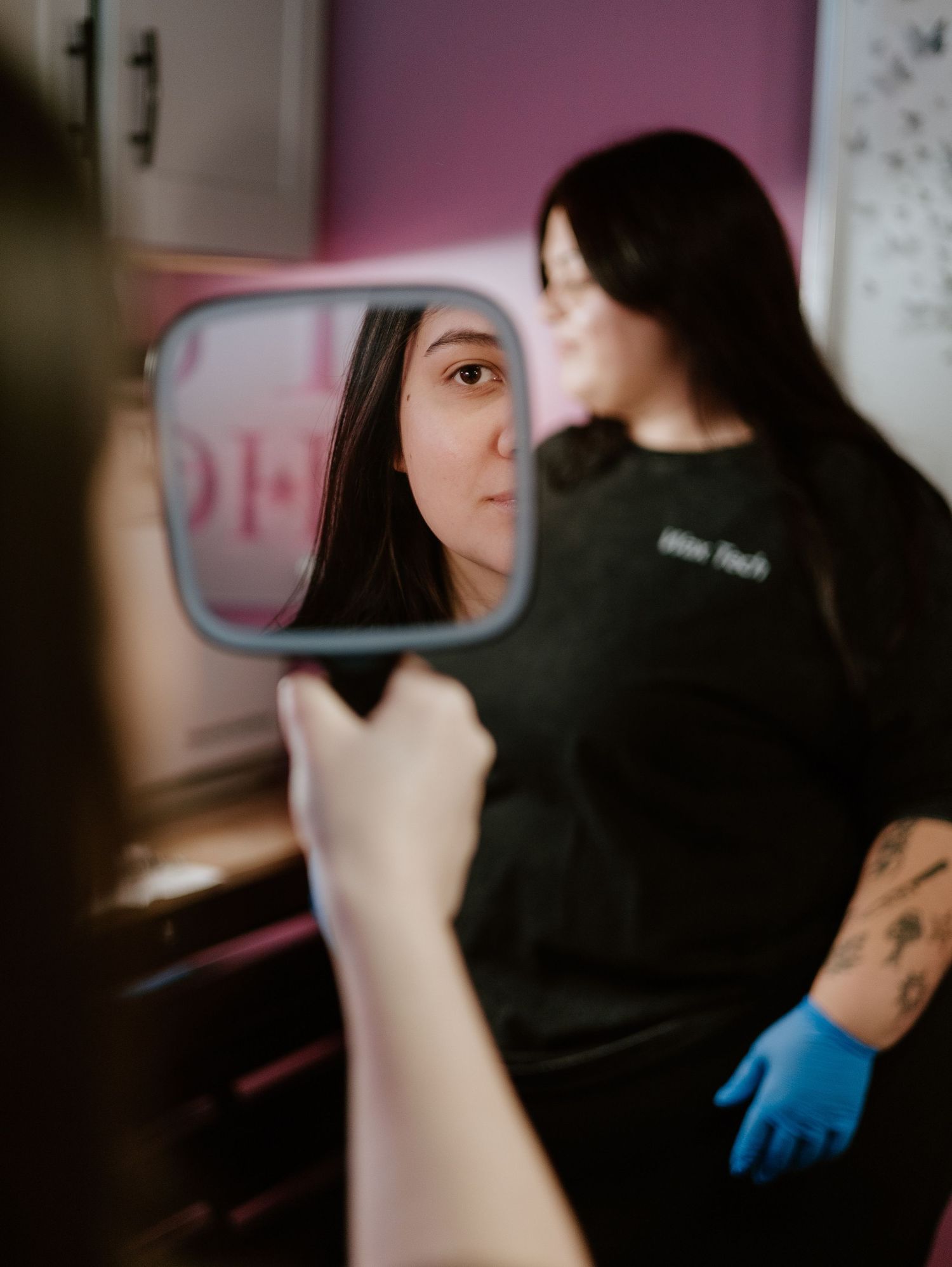Reflection in a small mirror shows someone in a black shirt while wearing blue gloves in a pink-walled room.