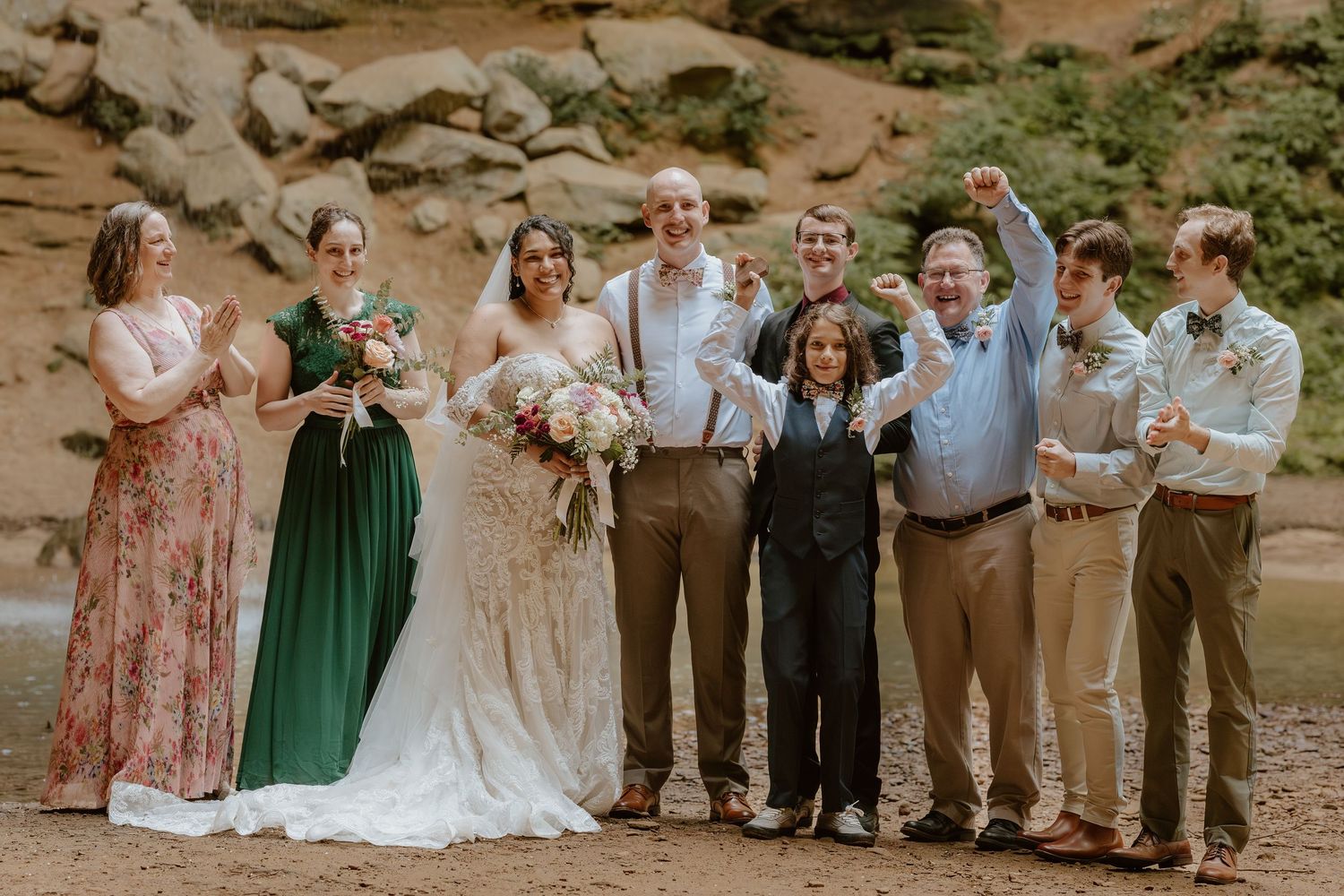 A wedding party poses together against a rocky outdoor backdrop wearing mixed formal attire.