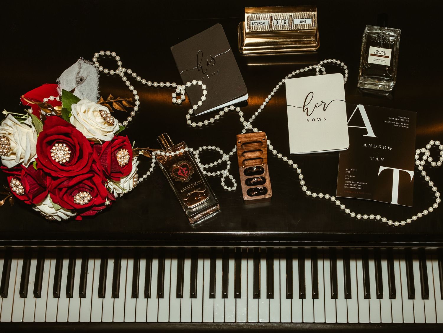 Wedding flat lay on piano keys with red rose bouquet, pearl necklace, perfume bottles, and monogram notebooks.