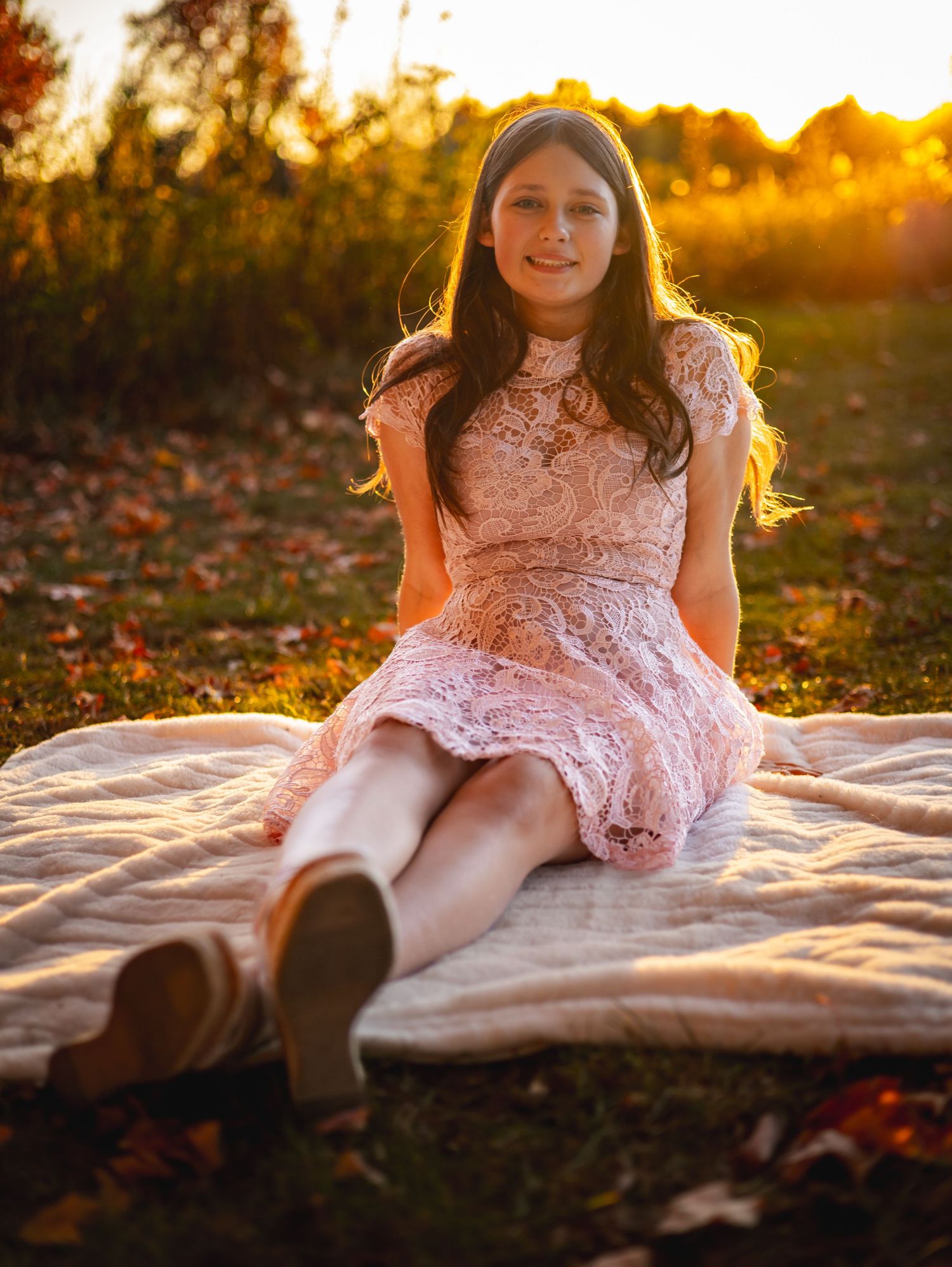 Someone in a pink lace dress sits on a blanket outdoors during golden hour sunset with fall leaves on the ground.