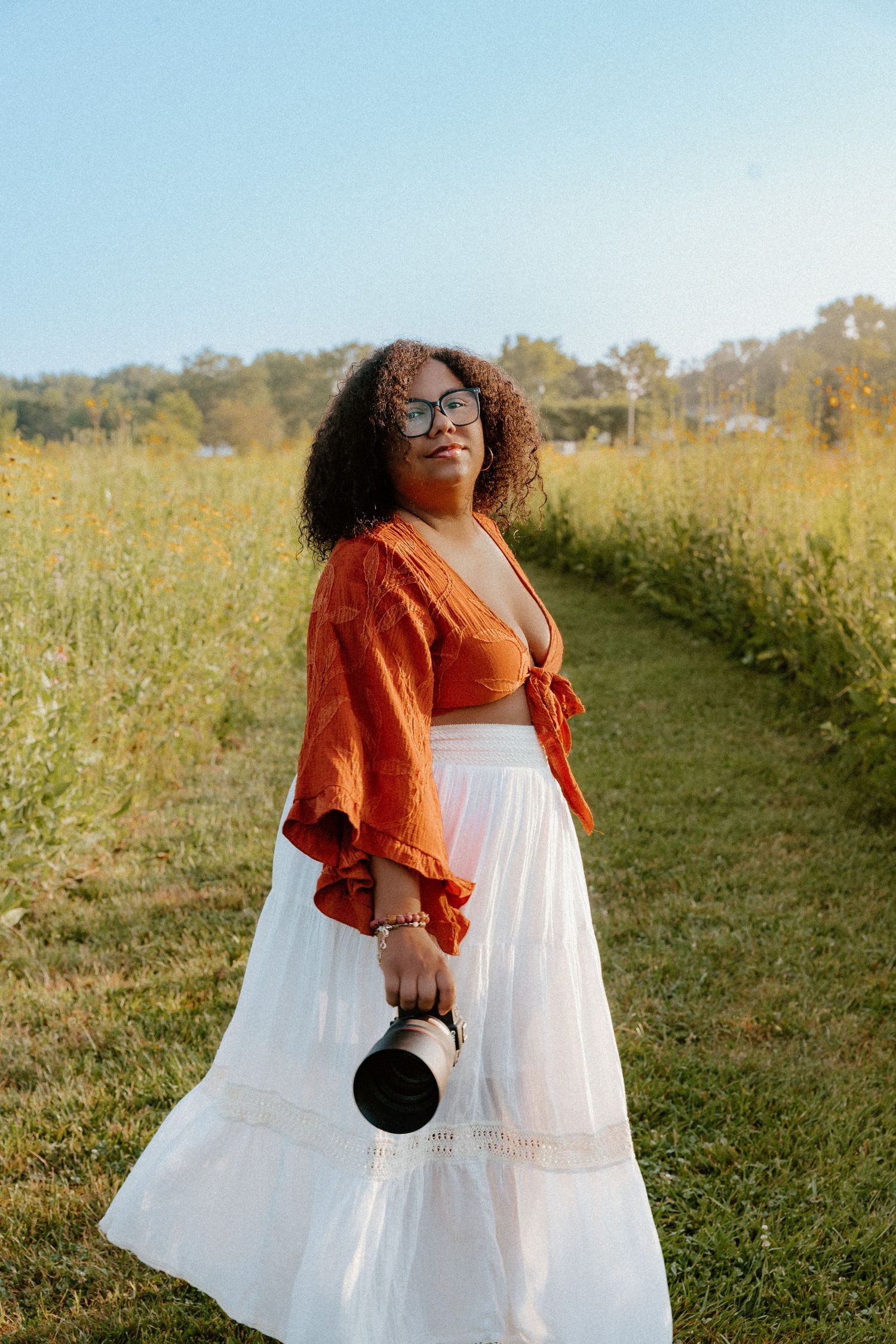 Person in white flowing dress and orange shrug stands in a sunlit meadow with golden wildflowers and trees in background.