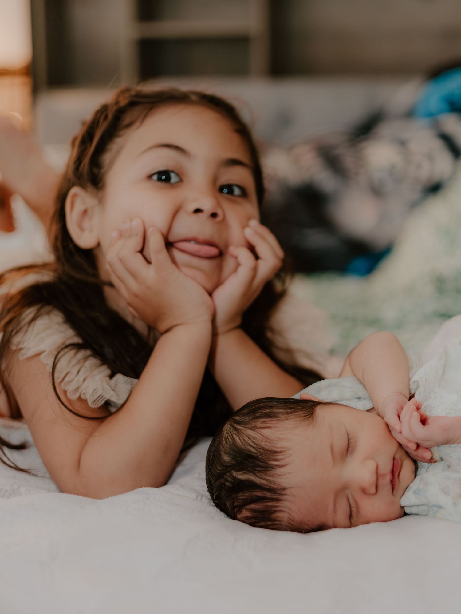 Two children lying on white bedding, with one awake resting head in hands while the other sleeps peacefully.