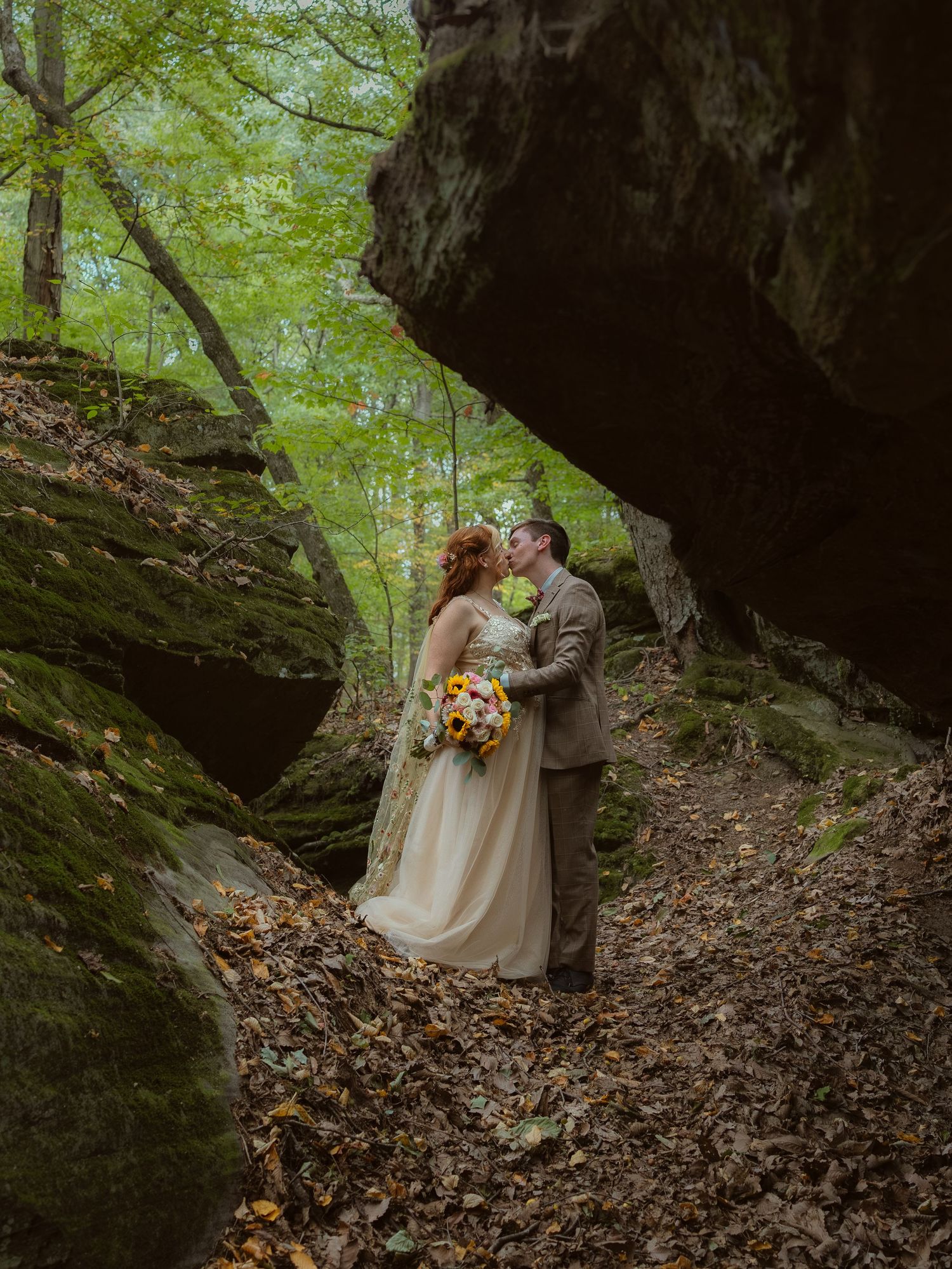 Wedding couple embraces near dramatic rock formations in a wooded forest setting with autumn leaves.