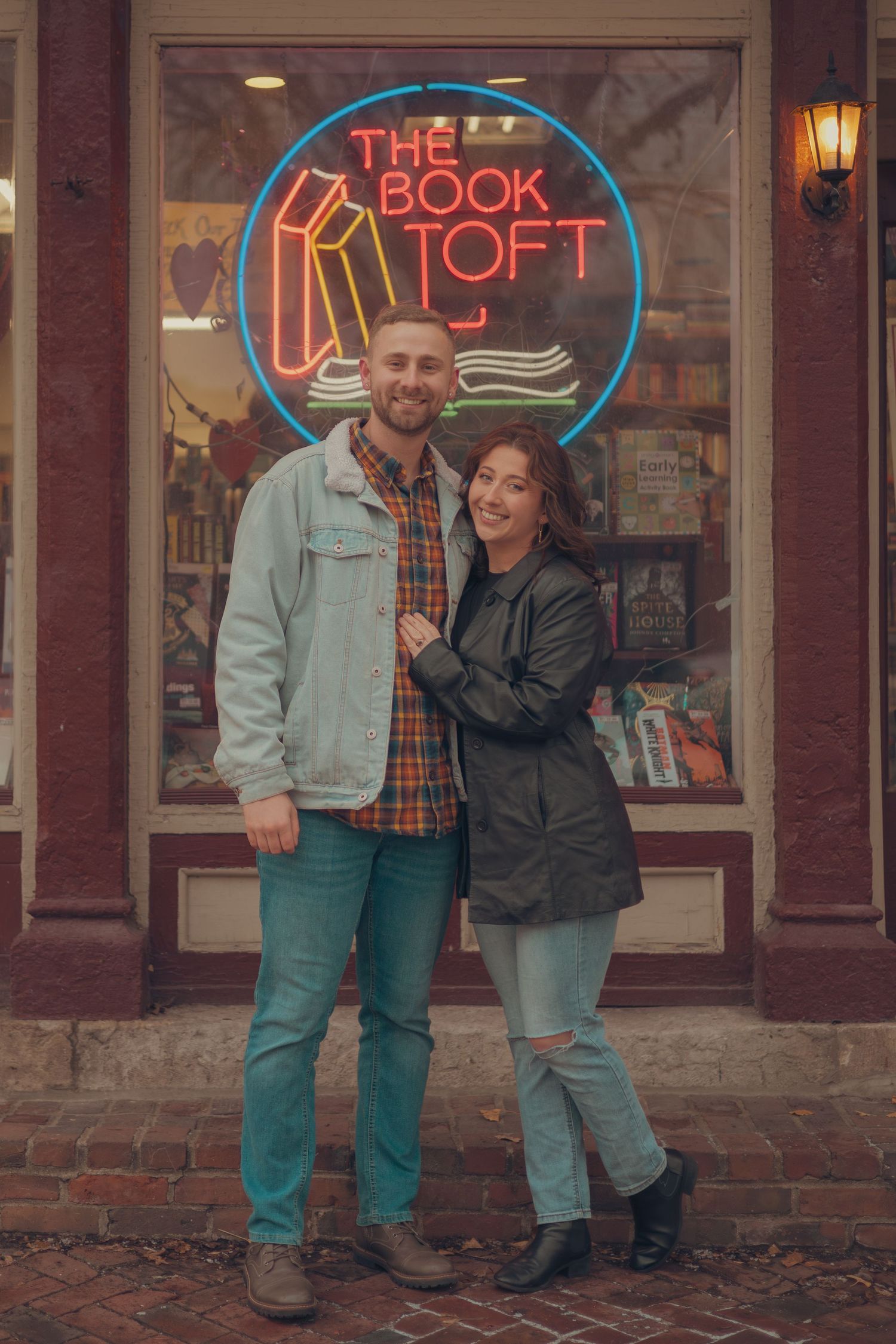 A couple poses outside a bookstore with neon signs and brick walls at dusk.