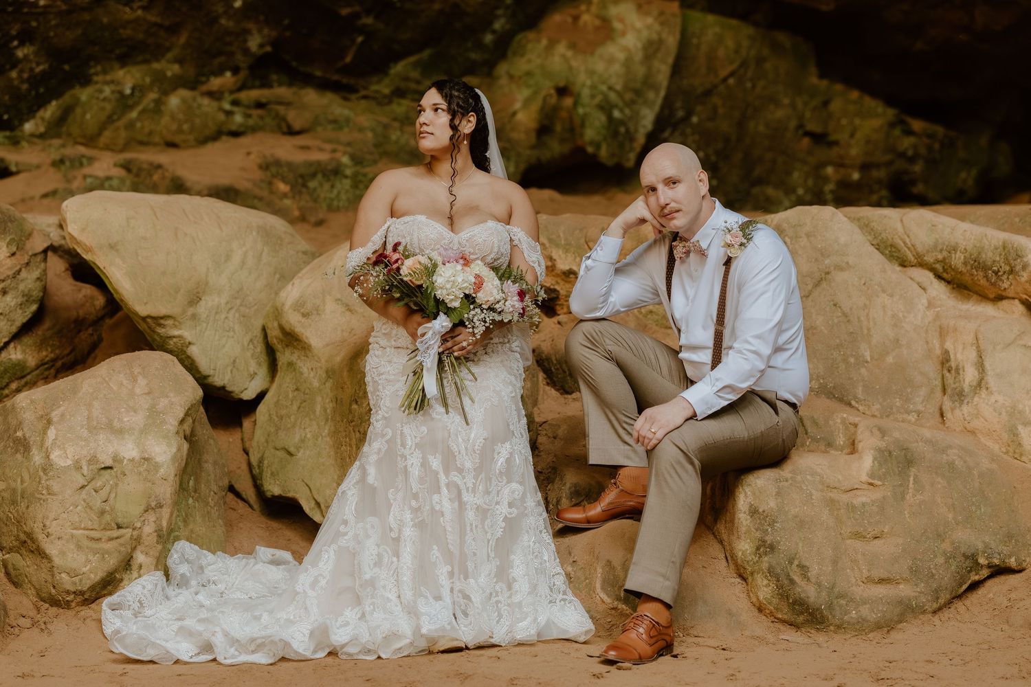 Newlyweds posing on large rocks outdoors with the bride holding a pink and white bouquet.