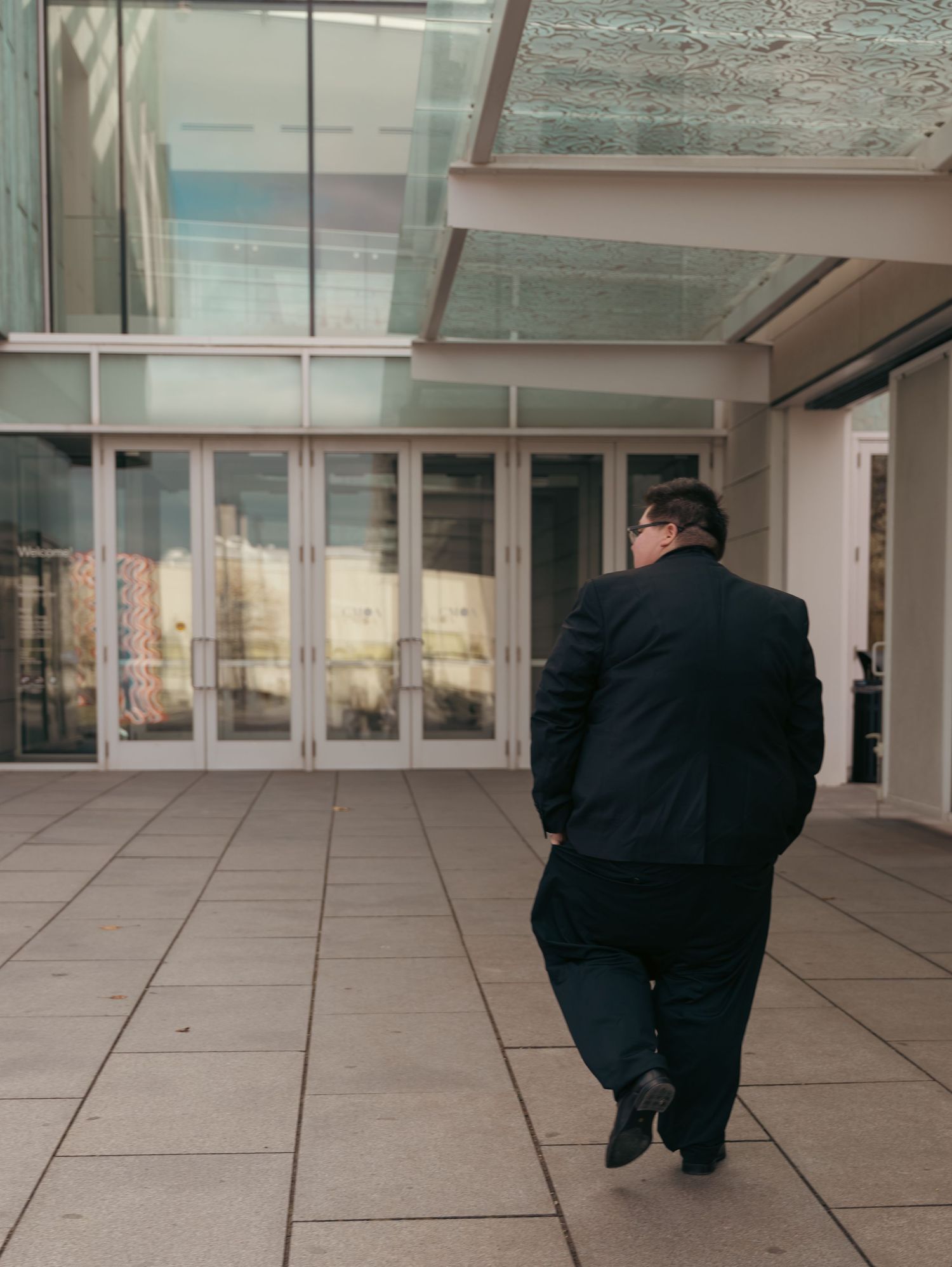 A figure in black clothing bends down in a modern gallery space with glass walls and tiled floor.
