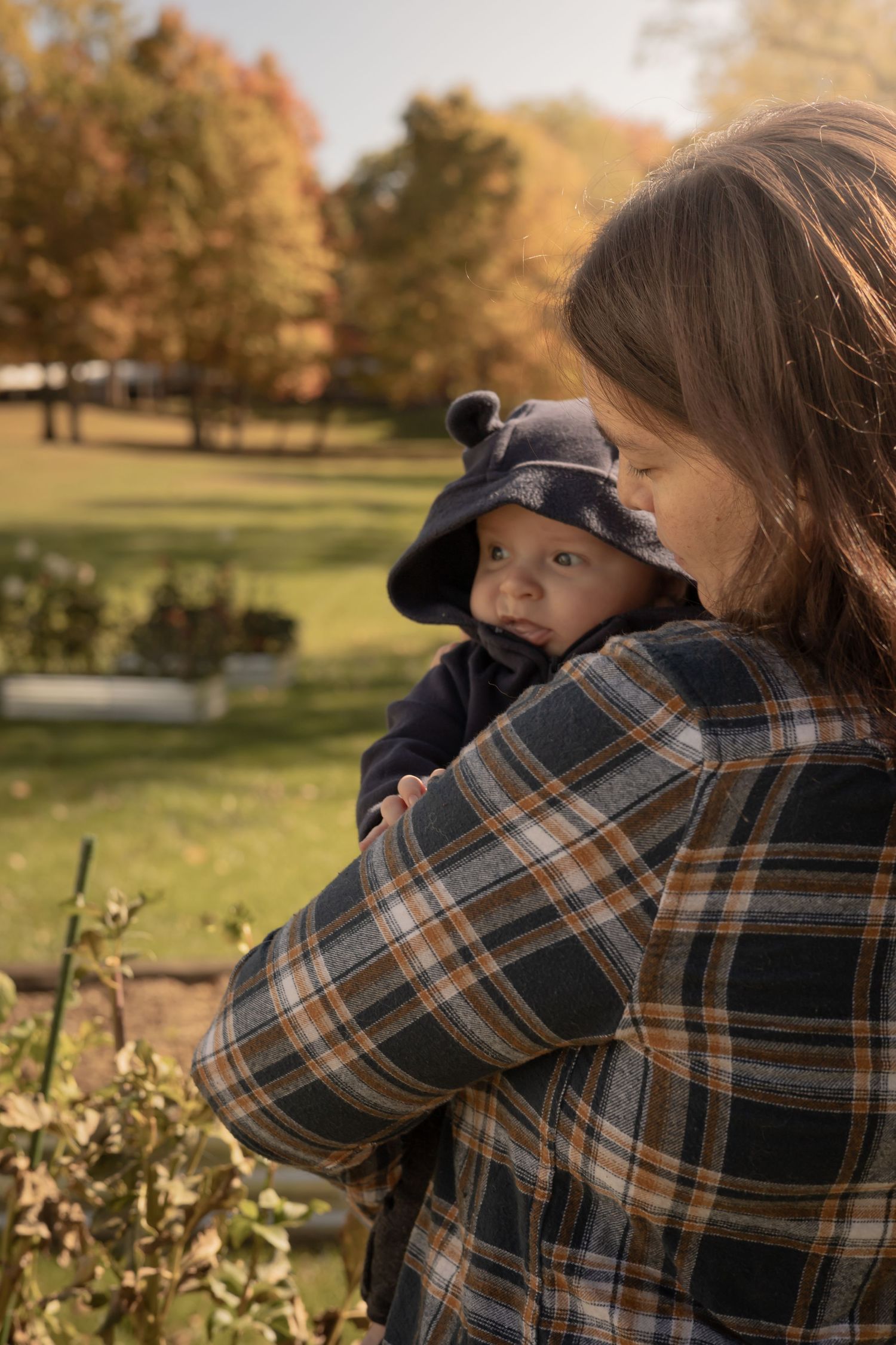 Person wearing a plaid flannel shirt holds a small baby in a blue hat outdoors during autumn with fall foliage in background.