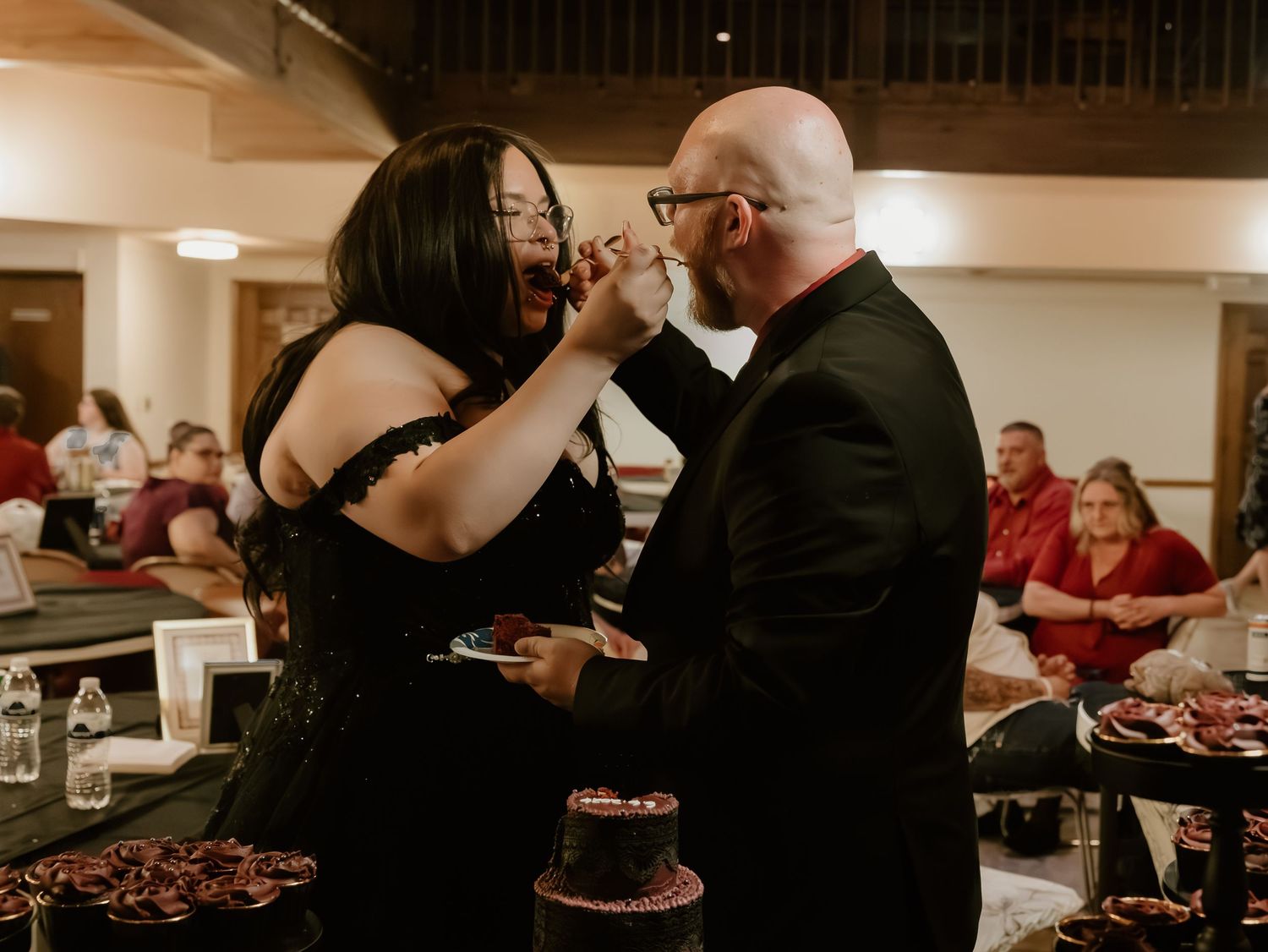 Couple sharing wedding cake at reception, bride in black gown feeding groom in suit, guests seated in background.