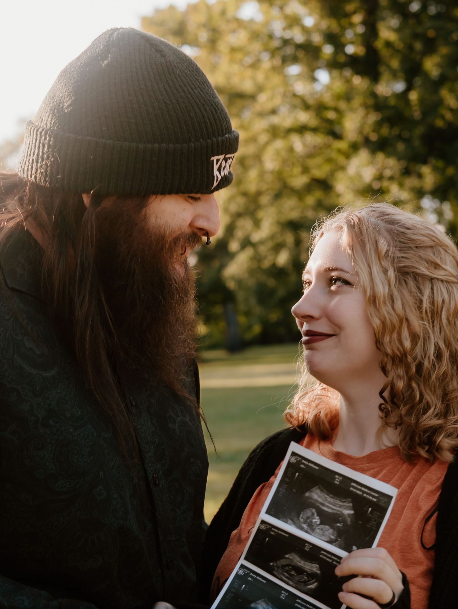 A couple shares an intimate moment outdoors while holding an ultrasound photo in autumn lighting.
