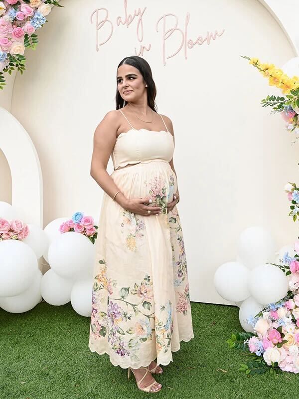 Expectant mother in floral cream dress posing at baby shower with pink and white balloon arch and flower decorations.