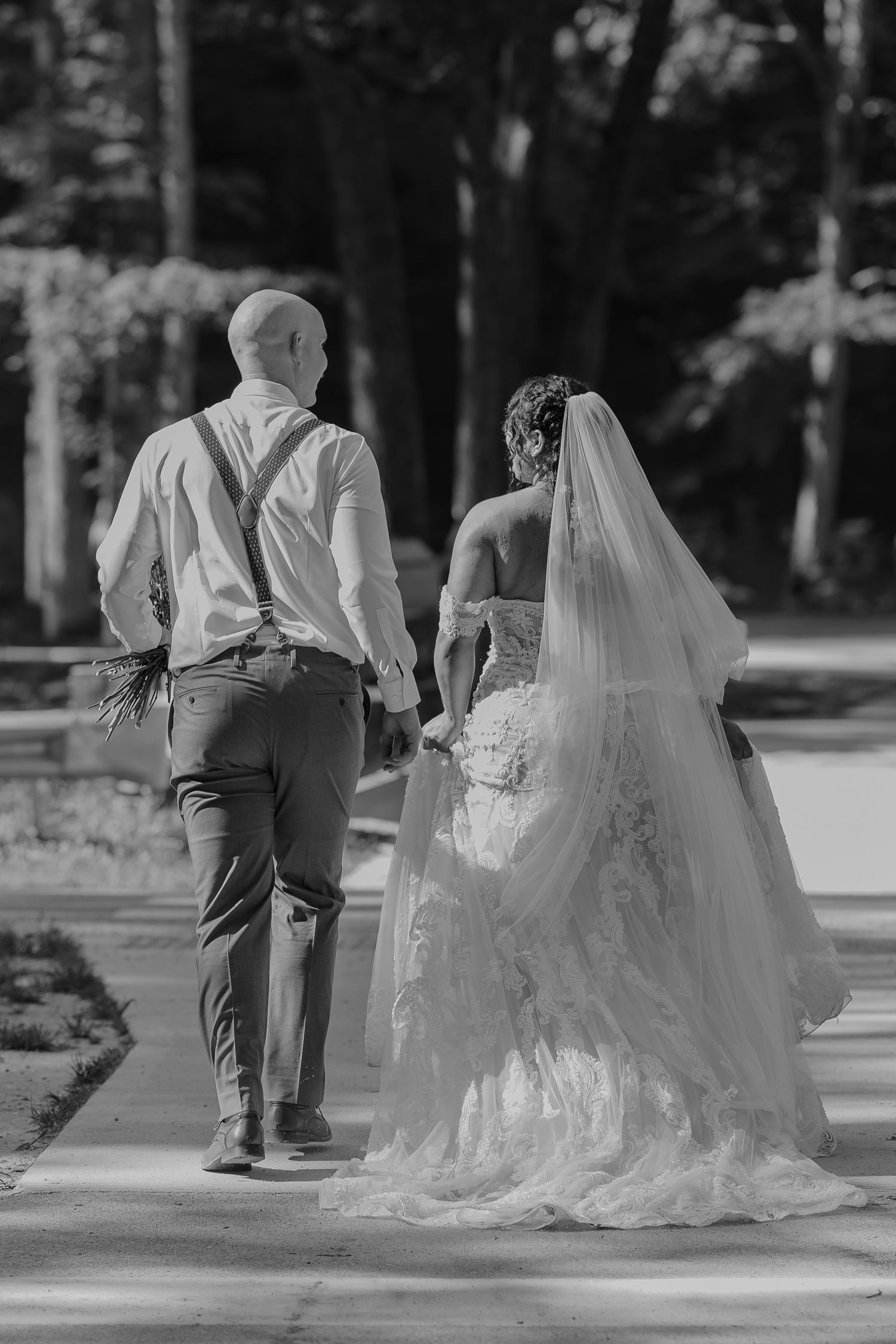 A black and white photo of a bride and groom walking together on a tree-lined path.