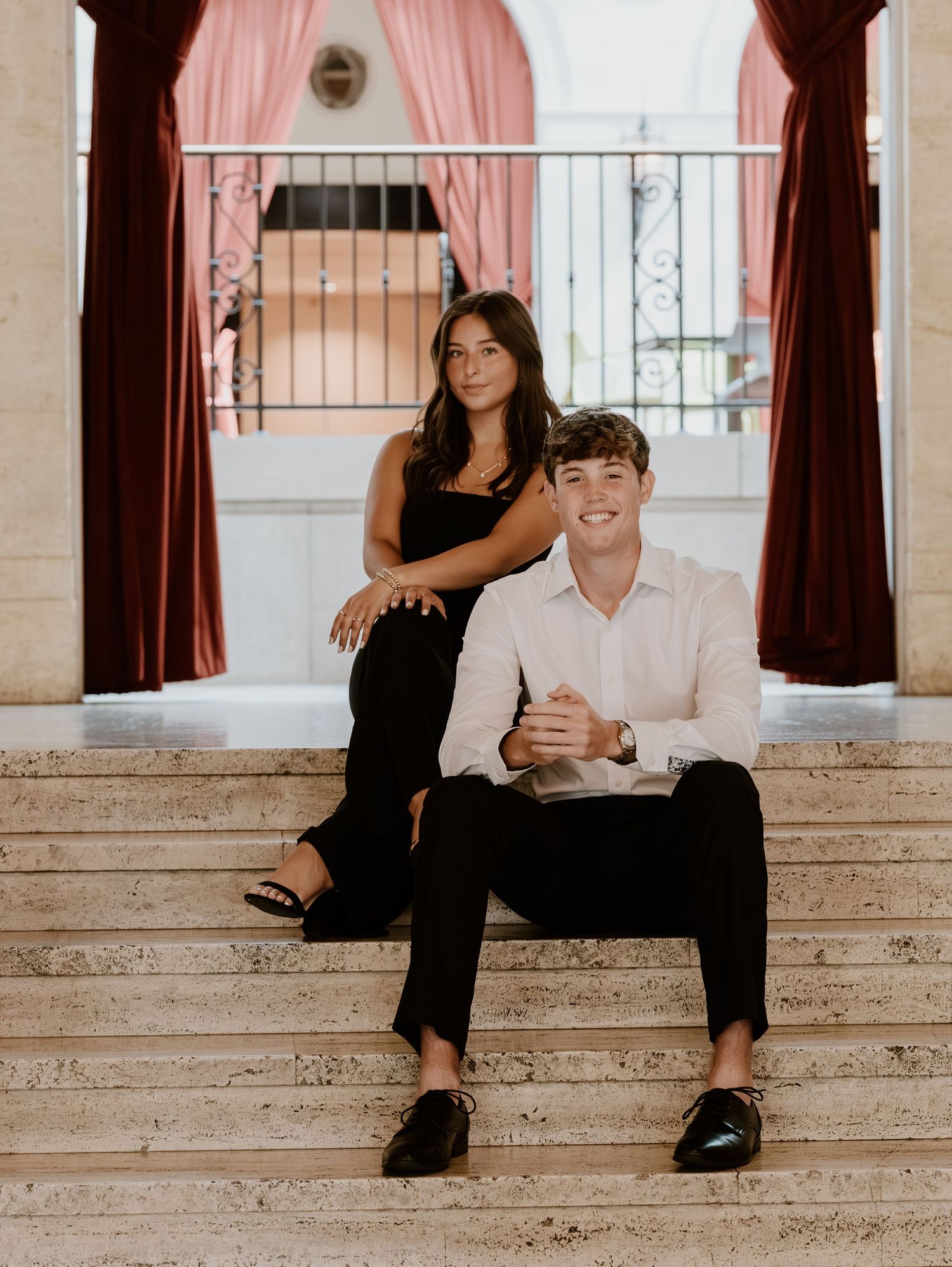 A couple relaxes on marble steps with burgundy curtains and a balcony railing in the background.
