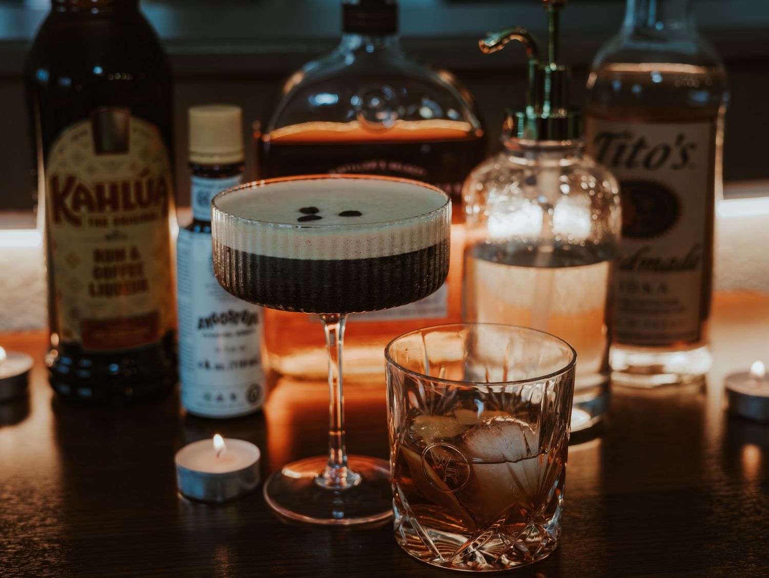 Close-up view of various whiskey bottles and cocktail glasses arranged on a dark wooden bar counter in dim lighting.