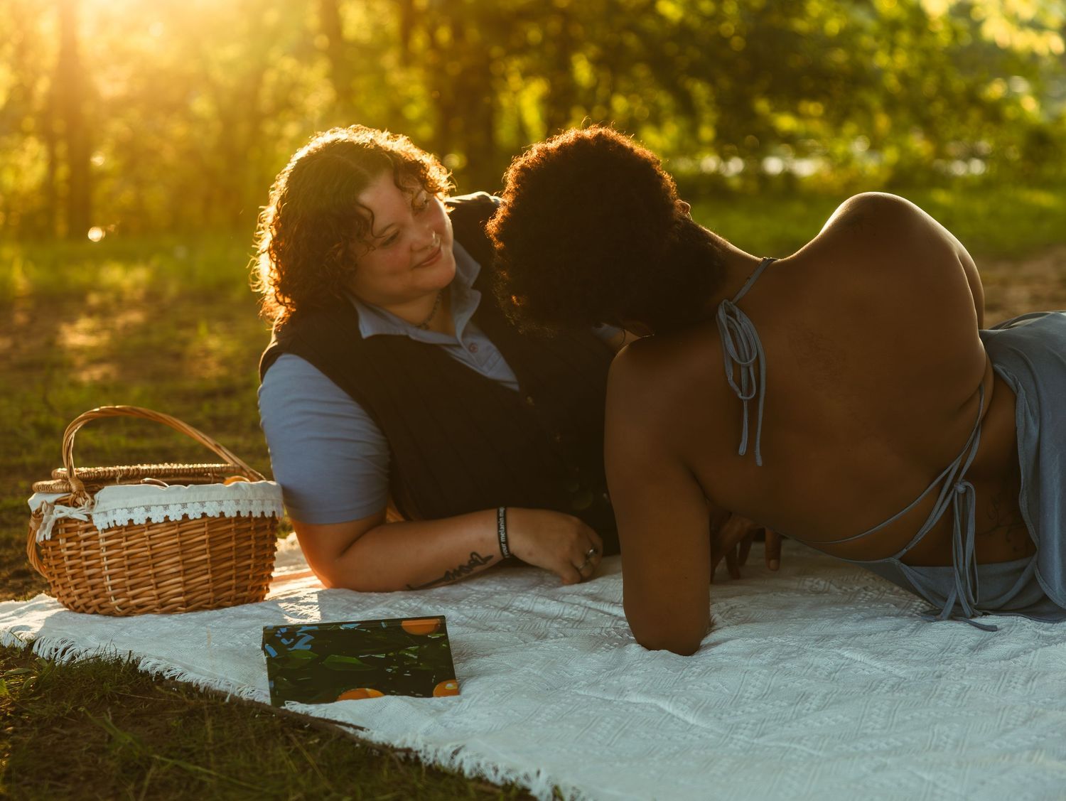 Two women share a romantic moment on a white blanket during a golden sunset picnic in a lush green park.