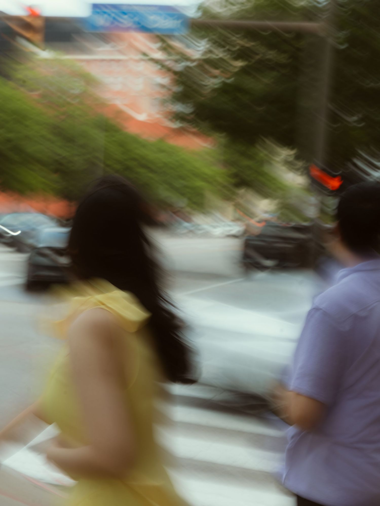 Blurry motion shot of a woman in yellow dress and man in blue shirt crossing a street in an urban area.