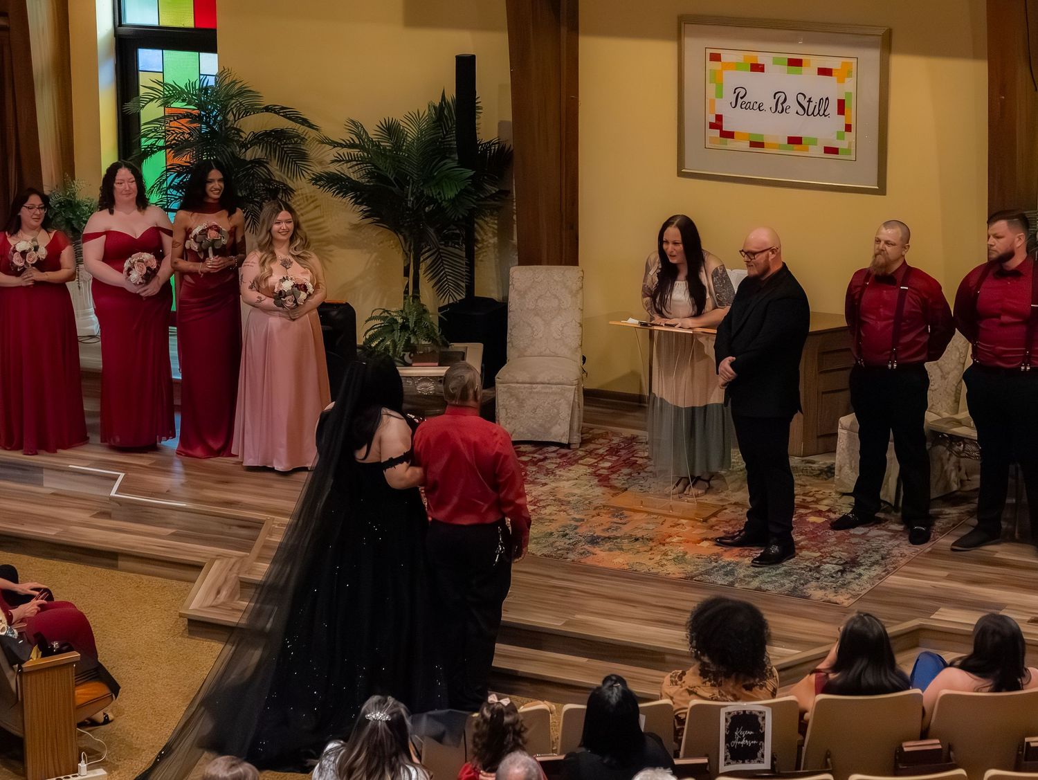 Wedding ceremony with bridesmaids in red and pink gowns, groomsmen in red shirts, as couple shares a kiss at the altar.