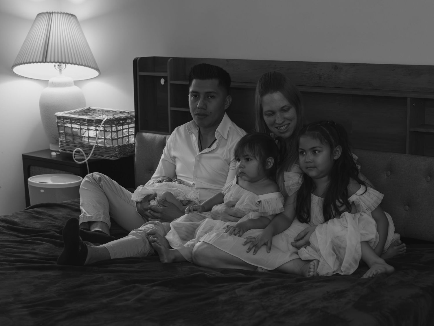 A family relaxes together on a bed under soft lamplight in black and white.