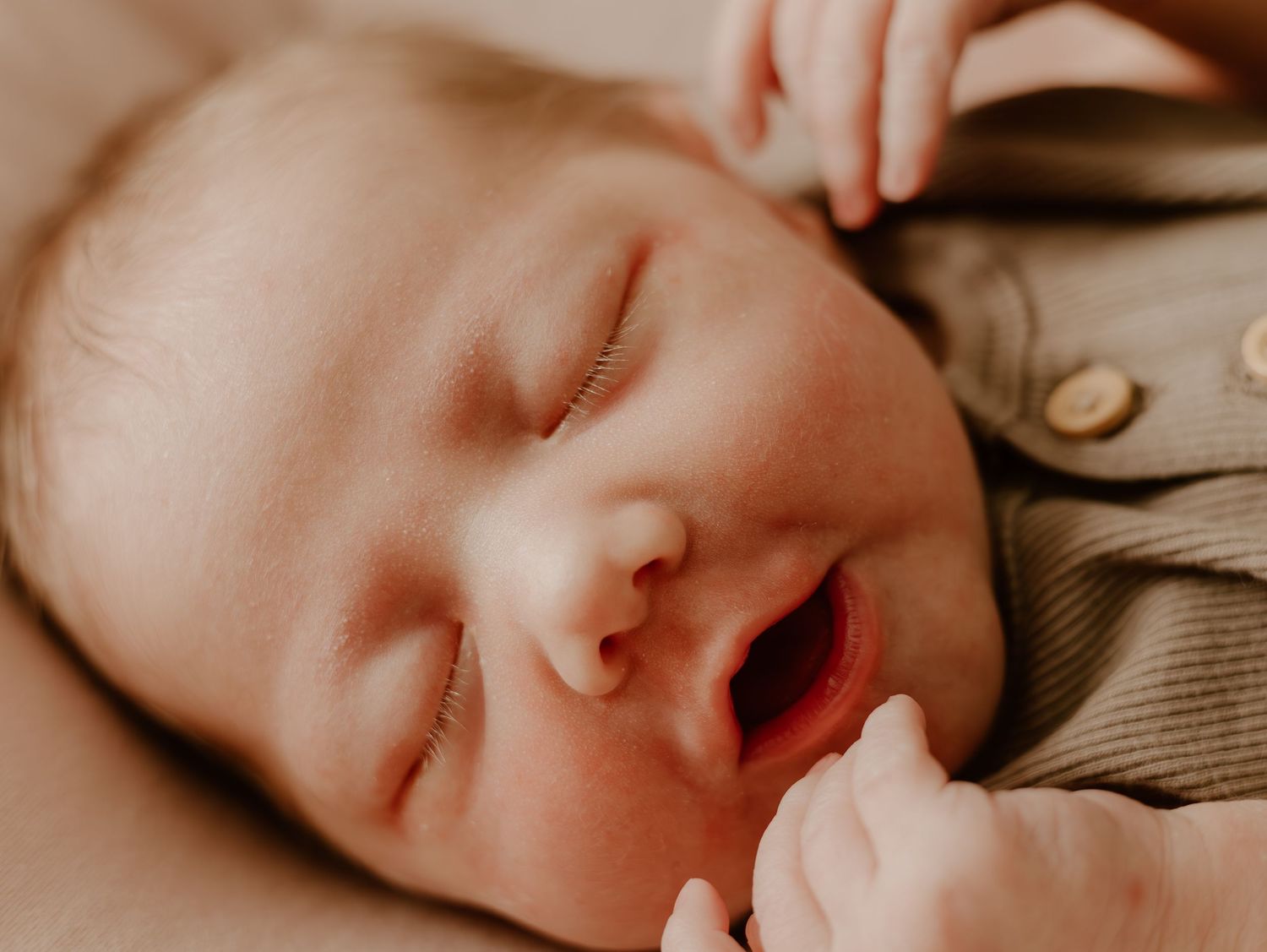 Close up view of a sleeping baby with slightly parted lips wearing a beige outfit in warm lighting.