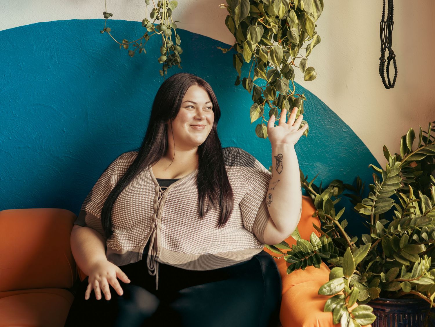 A woman in a beige sweater sits on a couch surrounded by plants and pumpkins in a cozy autumn setting.