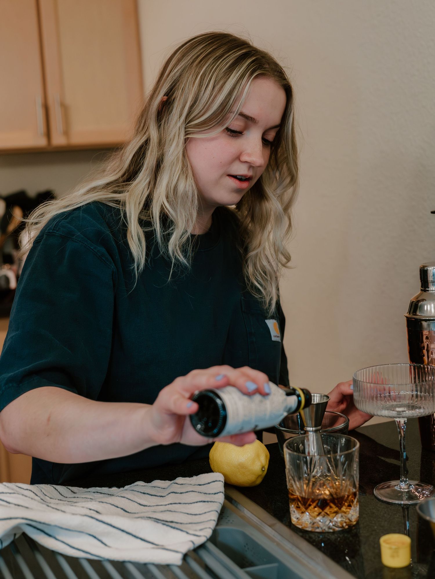 A person pours whiskey into a glass while preparing cocktails at a wooden table with citrus fruits and bar tools.