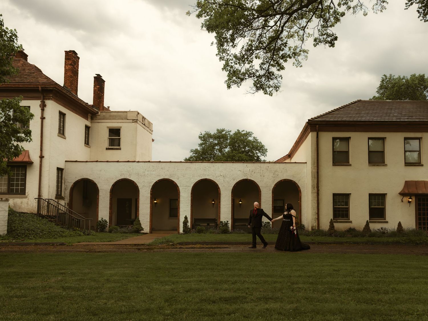 Couple in formal attire walking outside a historic white brick estate with arched colonnade and lush green lawn.