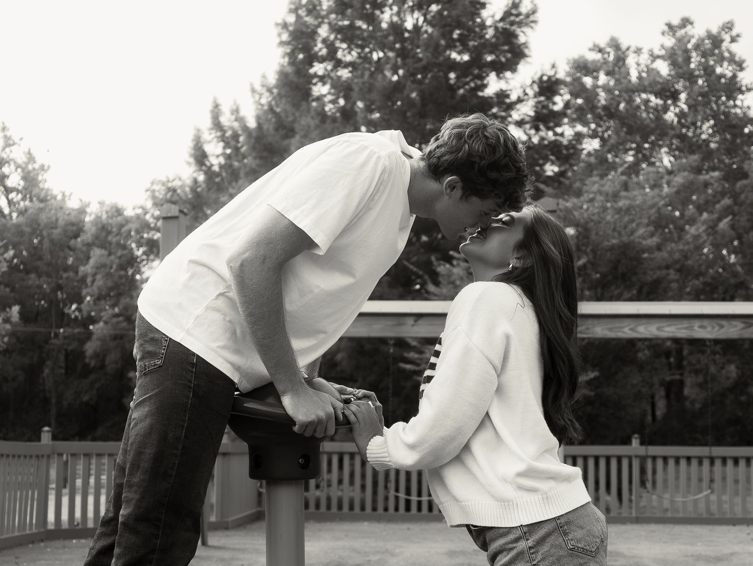 Black and white photo of a couple sharing a romantic kiss outdoors near a wooden deck surrounded by trees.