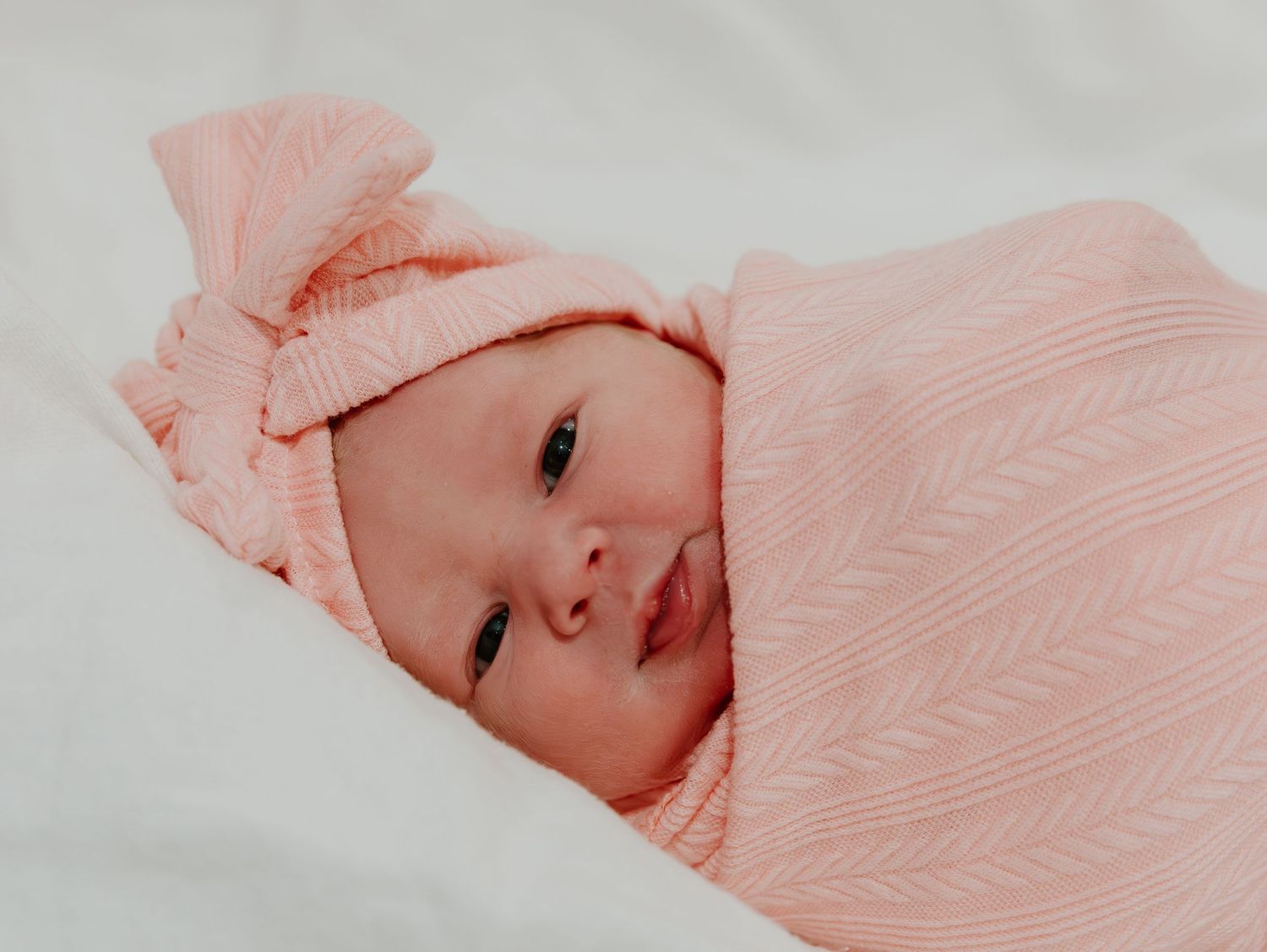 A tiny infant wrapped snugly in a soft pink knit blanket with a matching bow hat rests on white bedding.