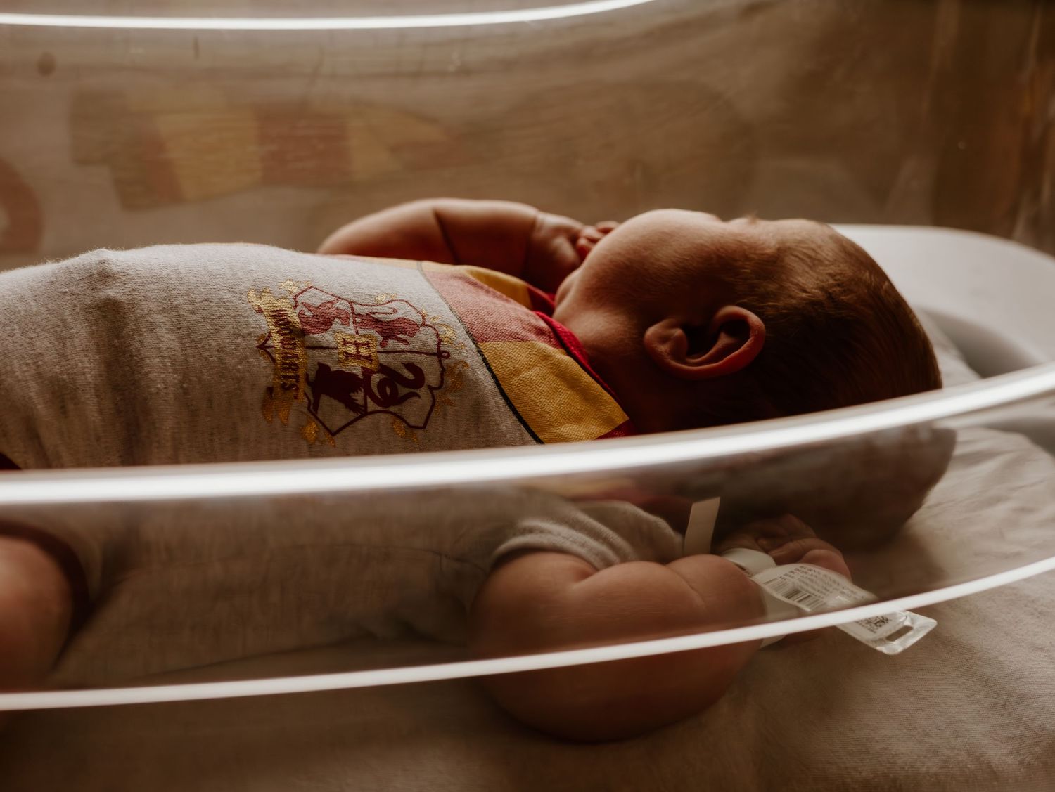 A newborn baby sleeps peacefully in a hospital bassinet wearing a brown swaddle blanket.