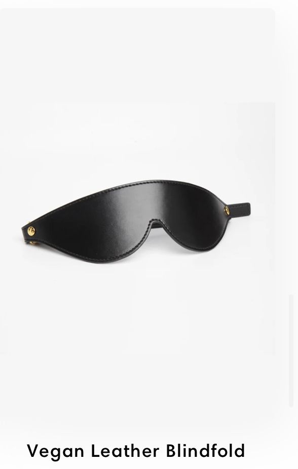A sleek black vegan leather blindfold with elastic strap and gold hardware against white background.