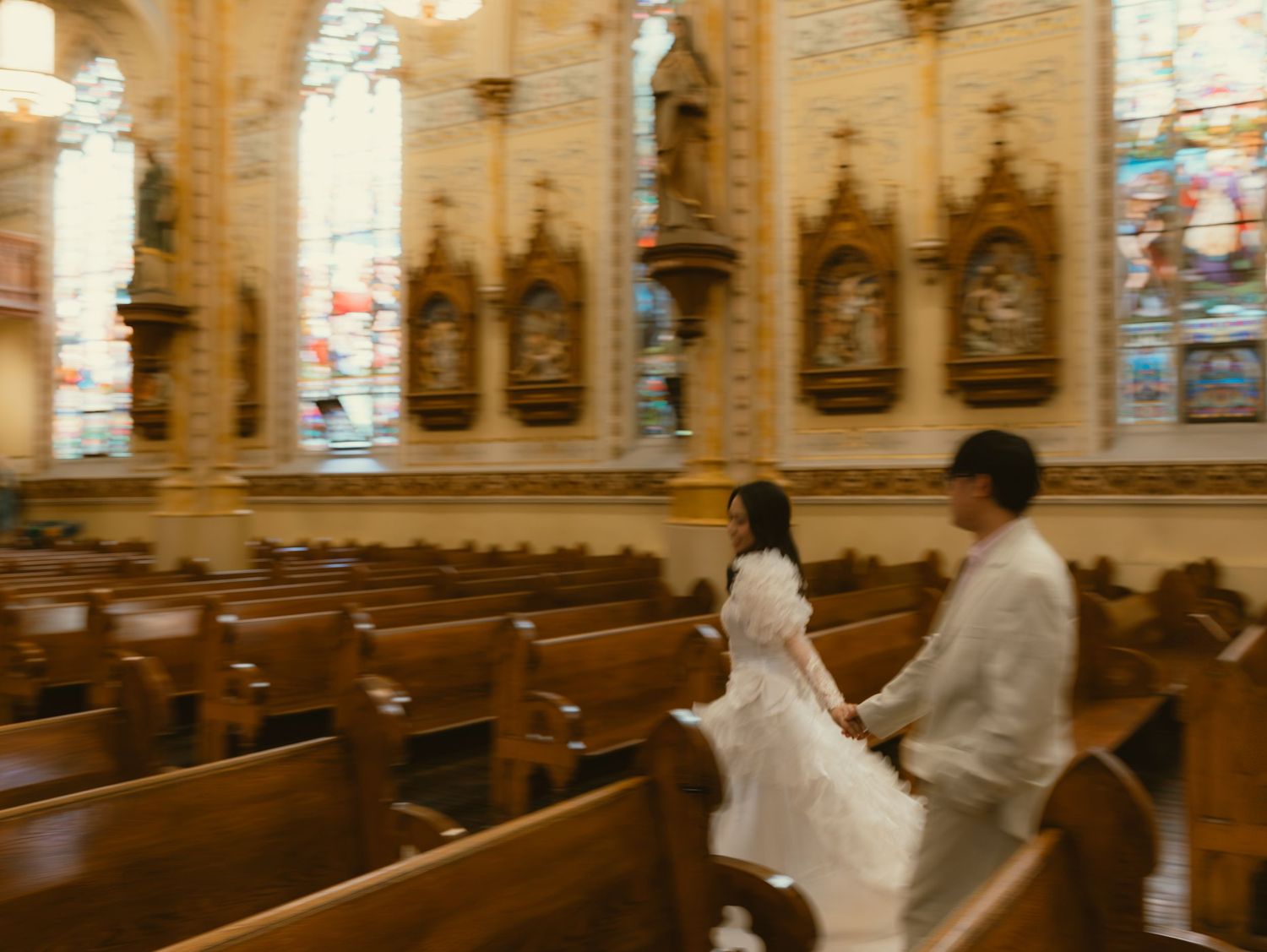 A couple in wedding attire walks hand-in-hand down the aisle of an ornate church with stained glass windows.