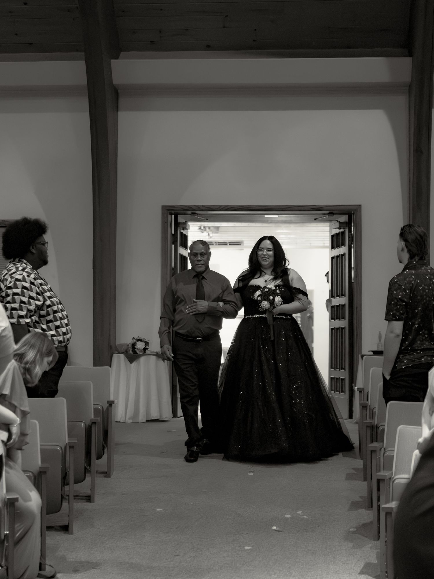 Black and white photo of a bride in a dark ball gown walking down the aisle with an escort at a wedding ceremony.