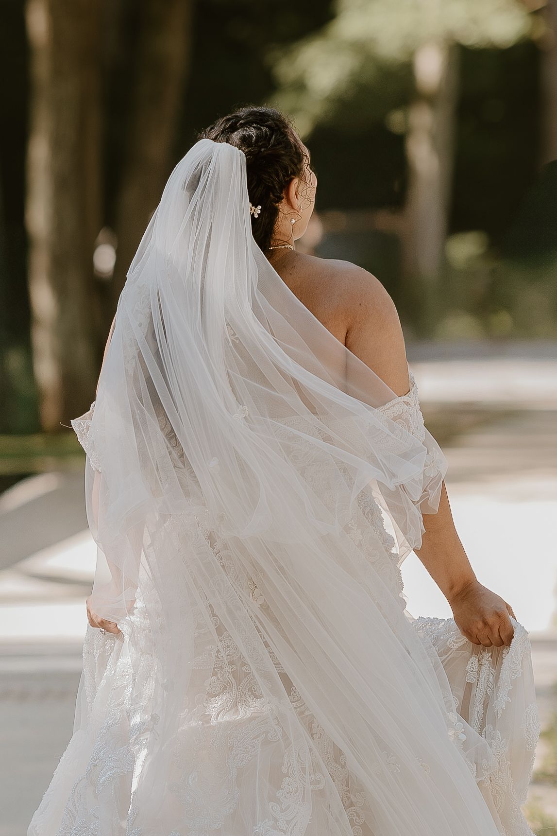 A flowing white wedding veil billows elegantly in the breeze as it trails behind a bride in an outdoor setting.