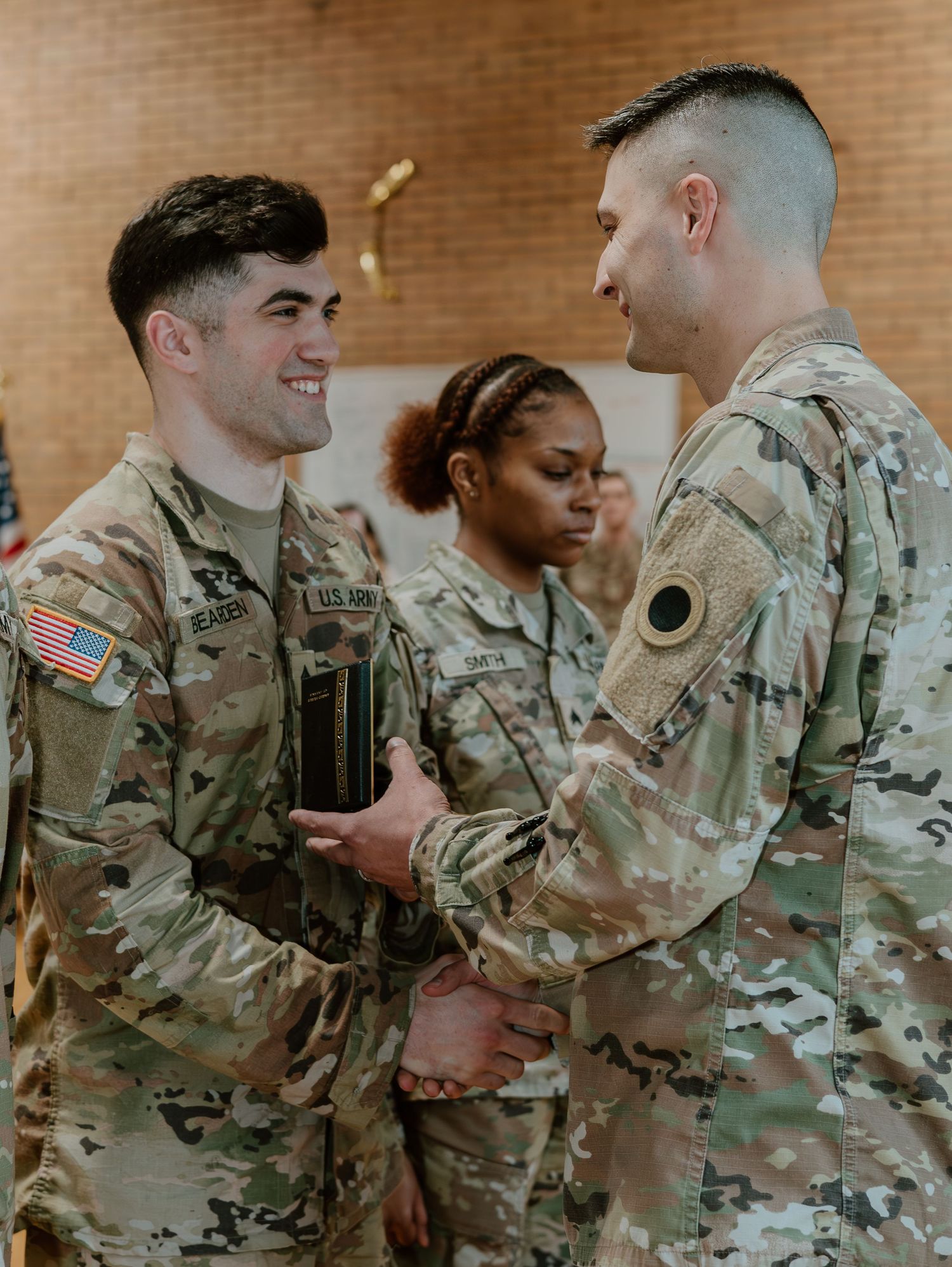 Military service members in camouflage uniforms share a warm interaction during what appears to be a ceremony or gathering.